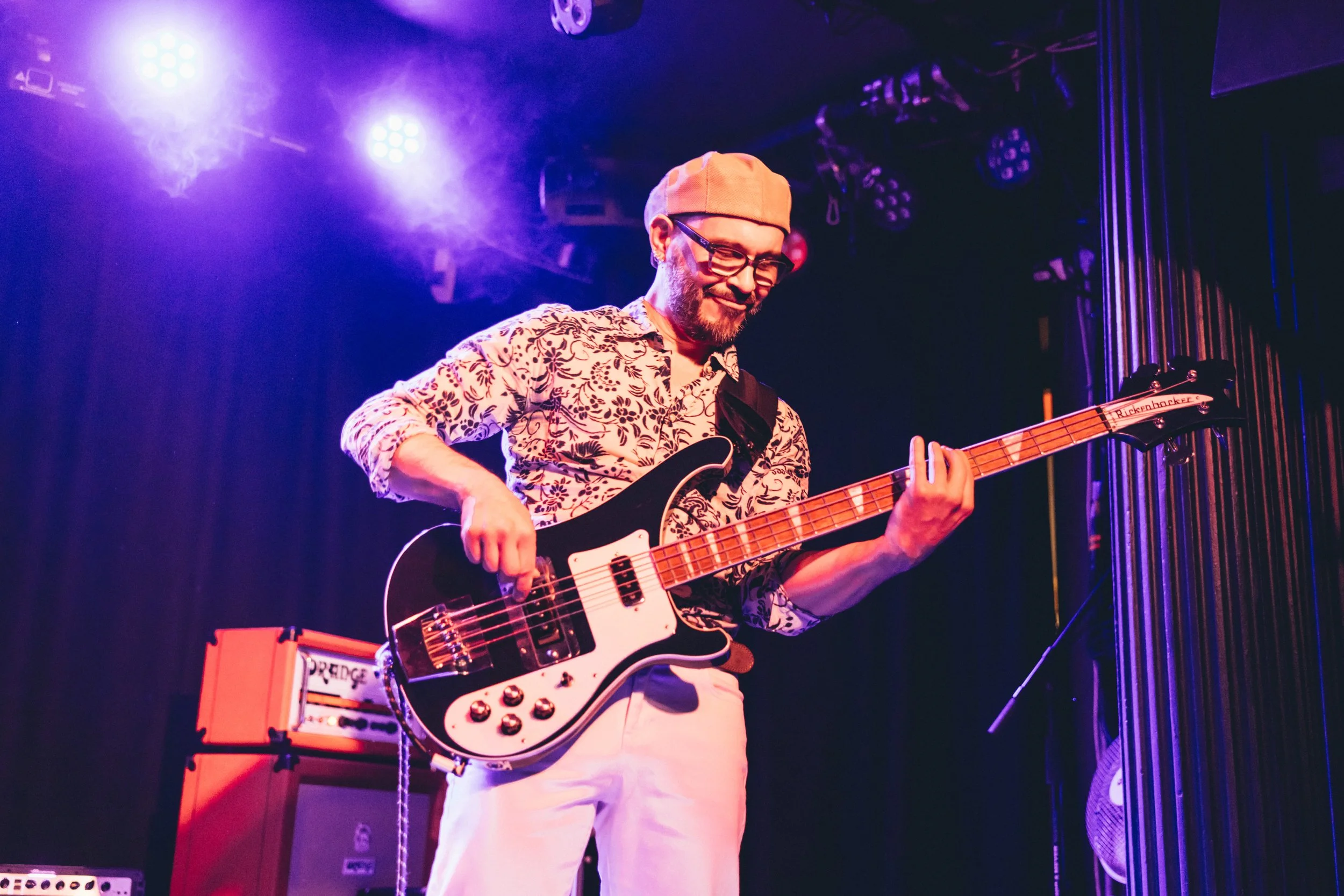 A man playing an electric bass guitar on stage under purple and blue stage lights, wearing a floral shirt, beige cap, glasses, and smiling.