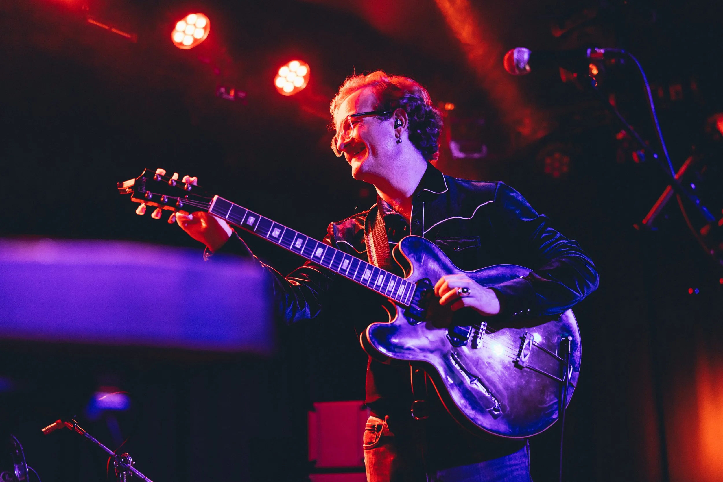 Person playing an electric guitar on stage with red and purple lighting.