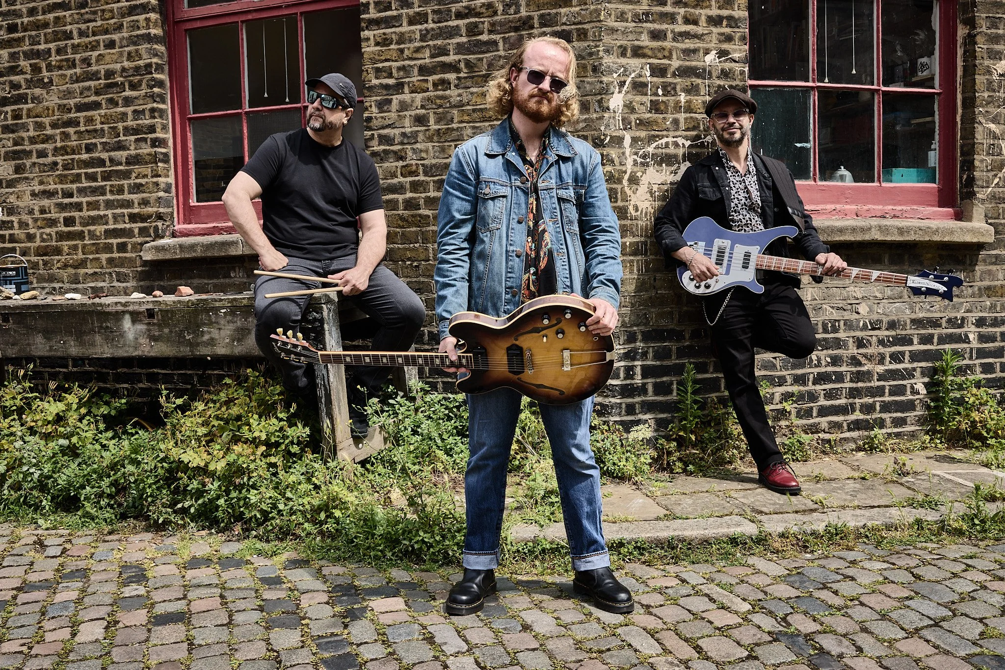 Three musicians outdoors on cobblestone street with brick wall and window in background. One man holds an electric guitar, one man holds an acoustic guitar, and the third man holds a bass guitar.
