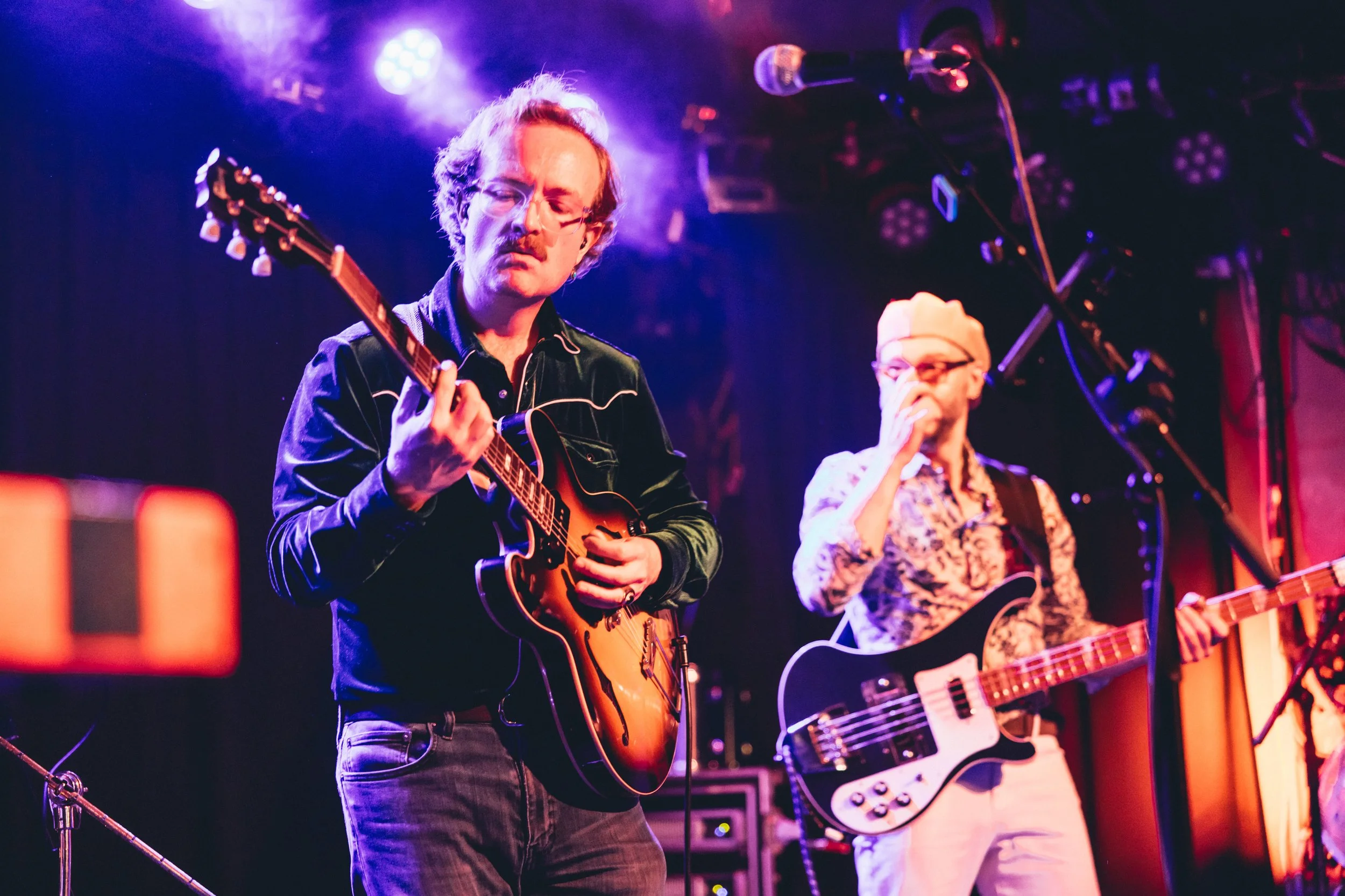Two musicians playing electric guitars on stage with colorful lighting.