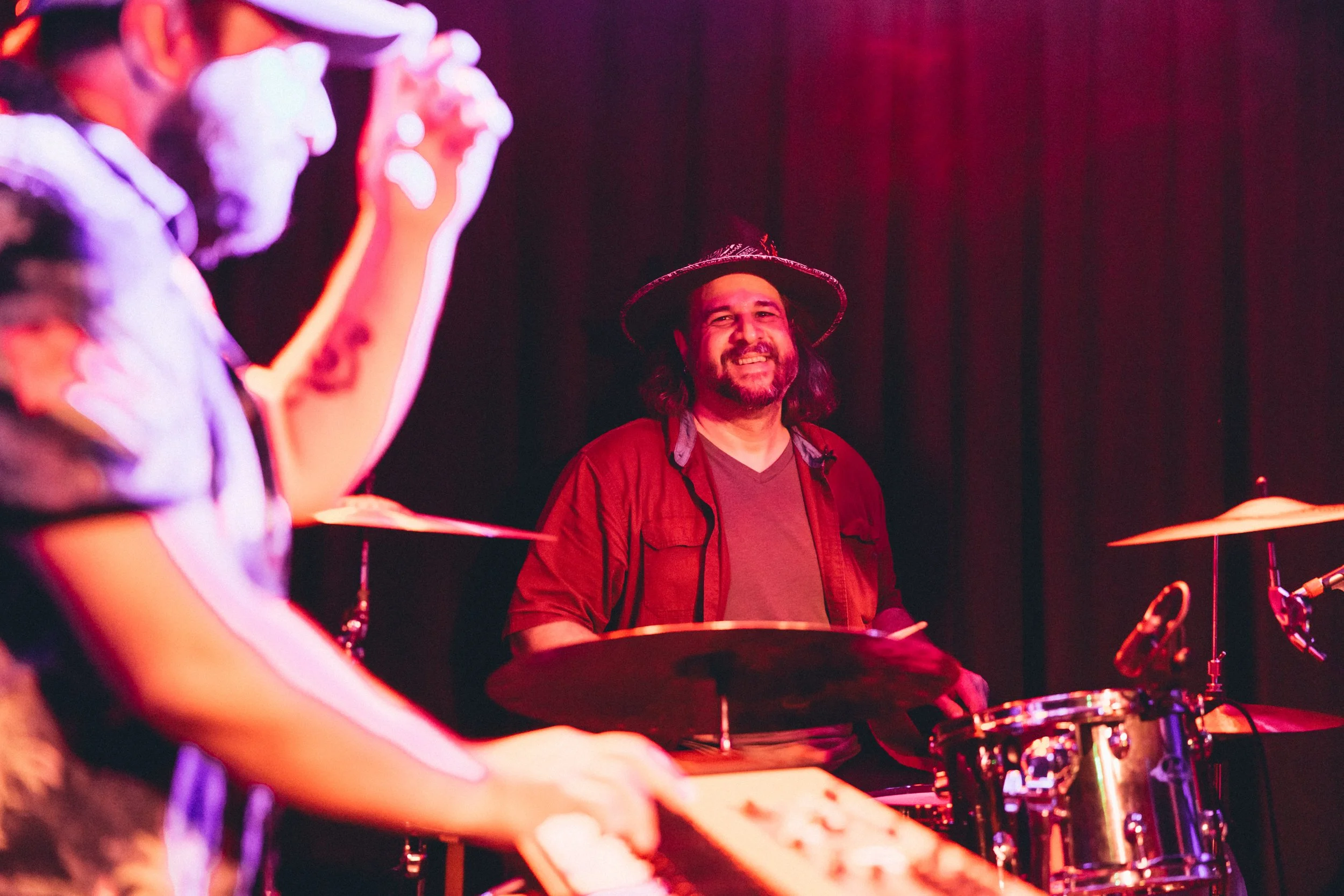 A man with long hair, beard, wearing a hat and red shirt, smiling while playing drums on stage with a dark background.