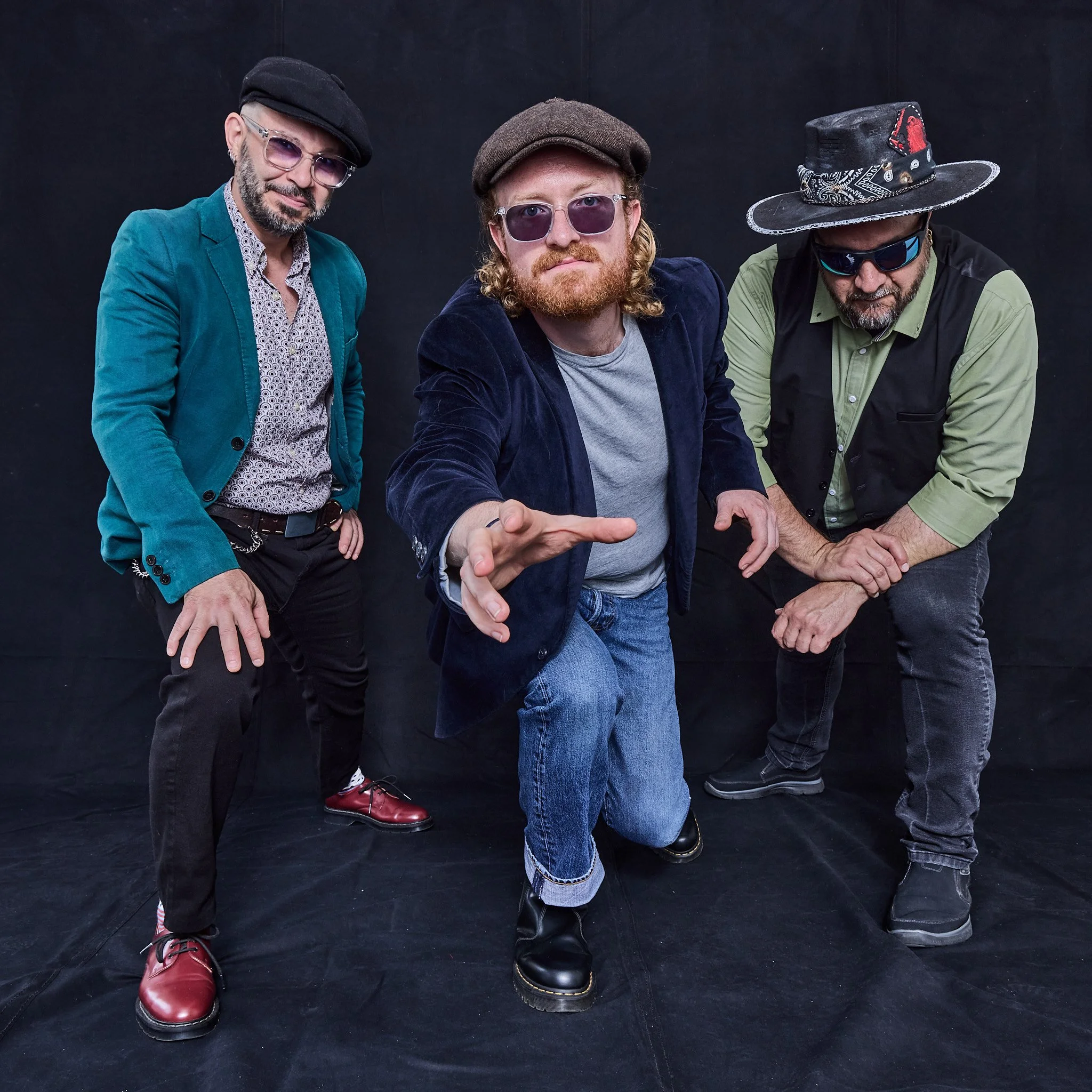 Three men dressed in vintage and retro clothing posing against a black background. One man is in front, reaching toward the camera. The others stand behind him, all wearing hats and sunglasses.