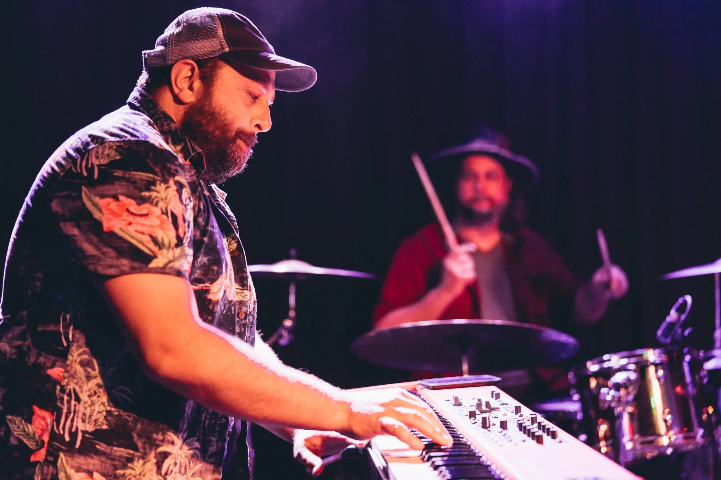 A man wearing a baseball cap and a vivid floral shirt playing a keyboard on stage, with a drummer in the background holding drumsticks and smiling.