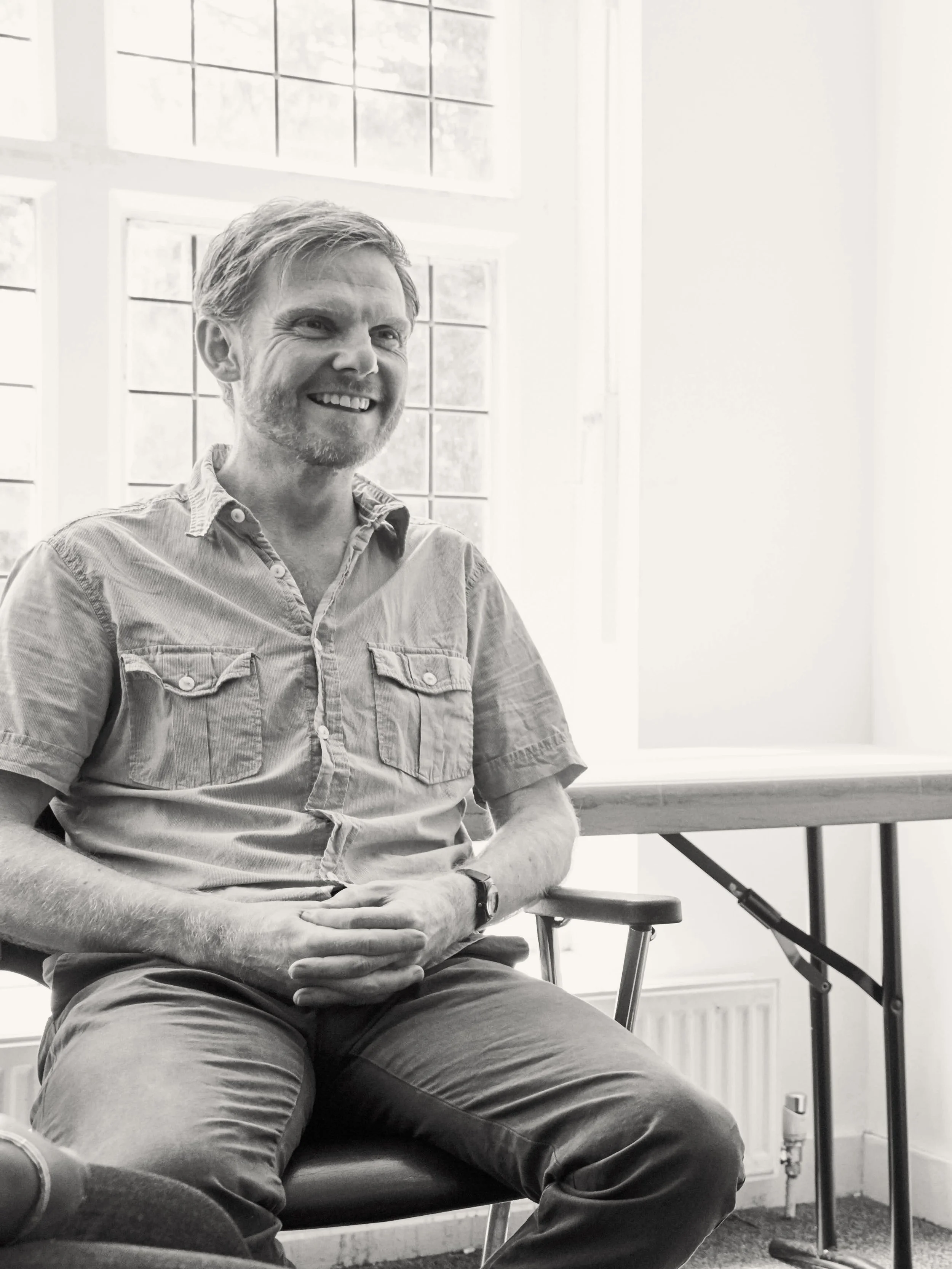 A smiling man with facial hair sitting in a chair, wearing a button-up shirt and pants inside a room with large windows.