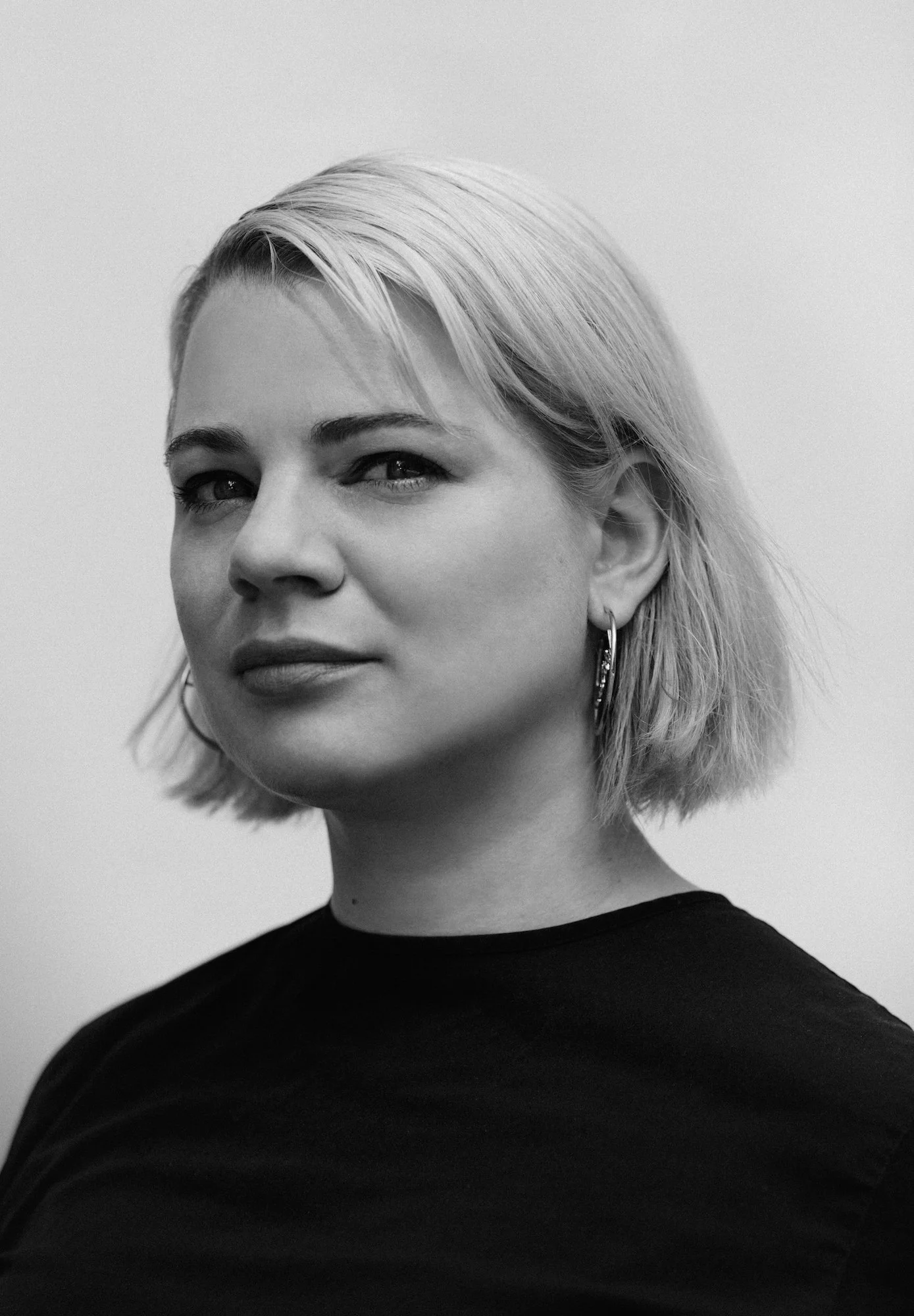 Black and white portrait of a woman with short blond hair, wearing hoop earrings and a black top, looking at the camera with a slight smile.