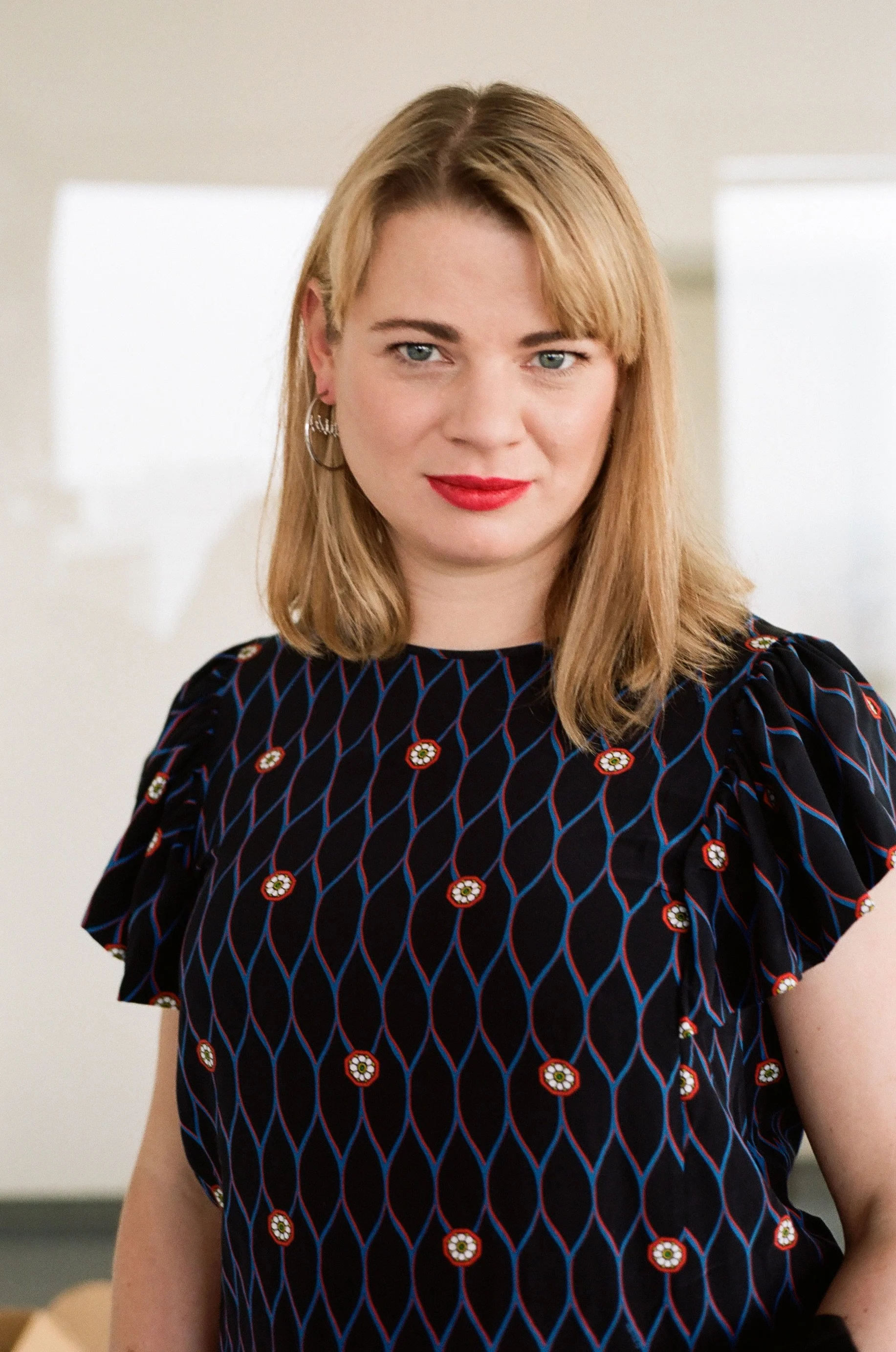A woman with shoulder-length blonde hair, wearing a dark patterned top, and red lipstick, standing indoors with a neutral background.