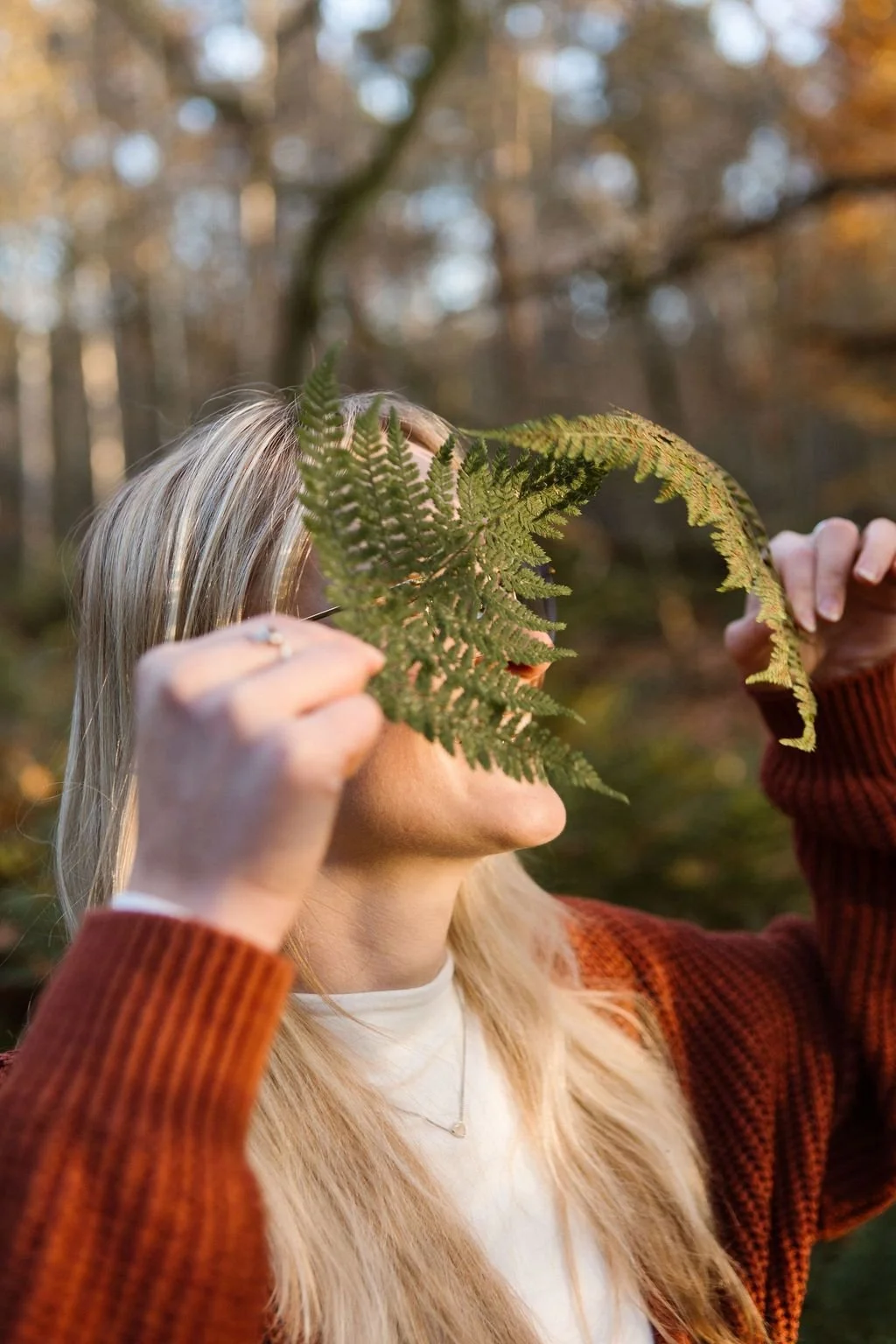 Vrouw met blonde haar houdt twee grote bladeren voor haar gezicht in een bos met herfstkleuren.