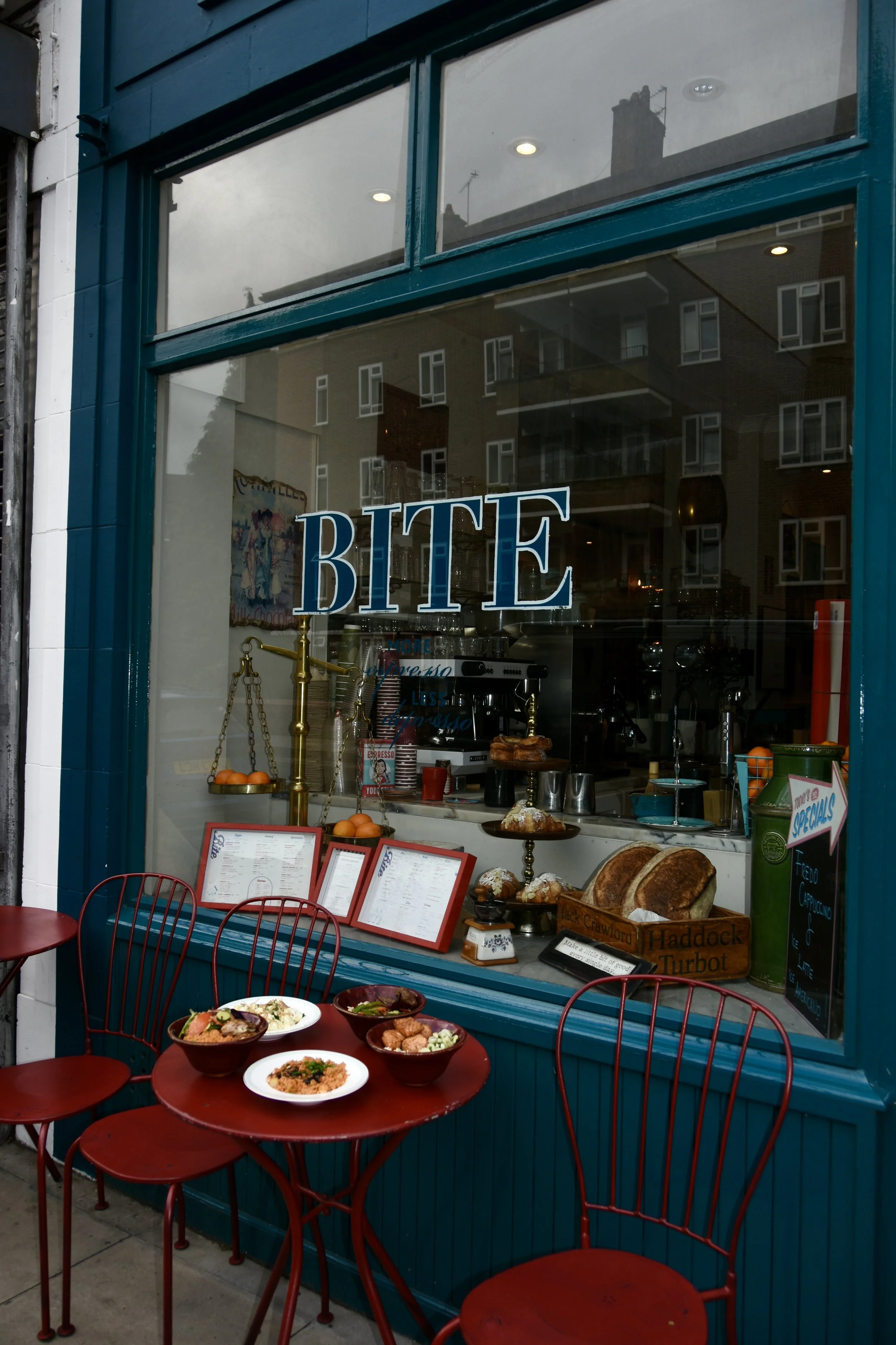 The image shows a storefront with a large window and teal trim, with the word 'BITE' in large letters on the window. Inside, there are baked goods, kitchen equipment, and a poster. Outside, there is a red table with four matching red chairs, set with bowls of food, on a sidewalk.