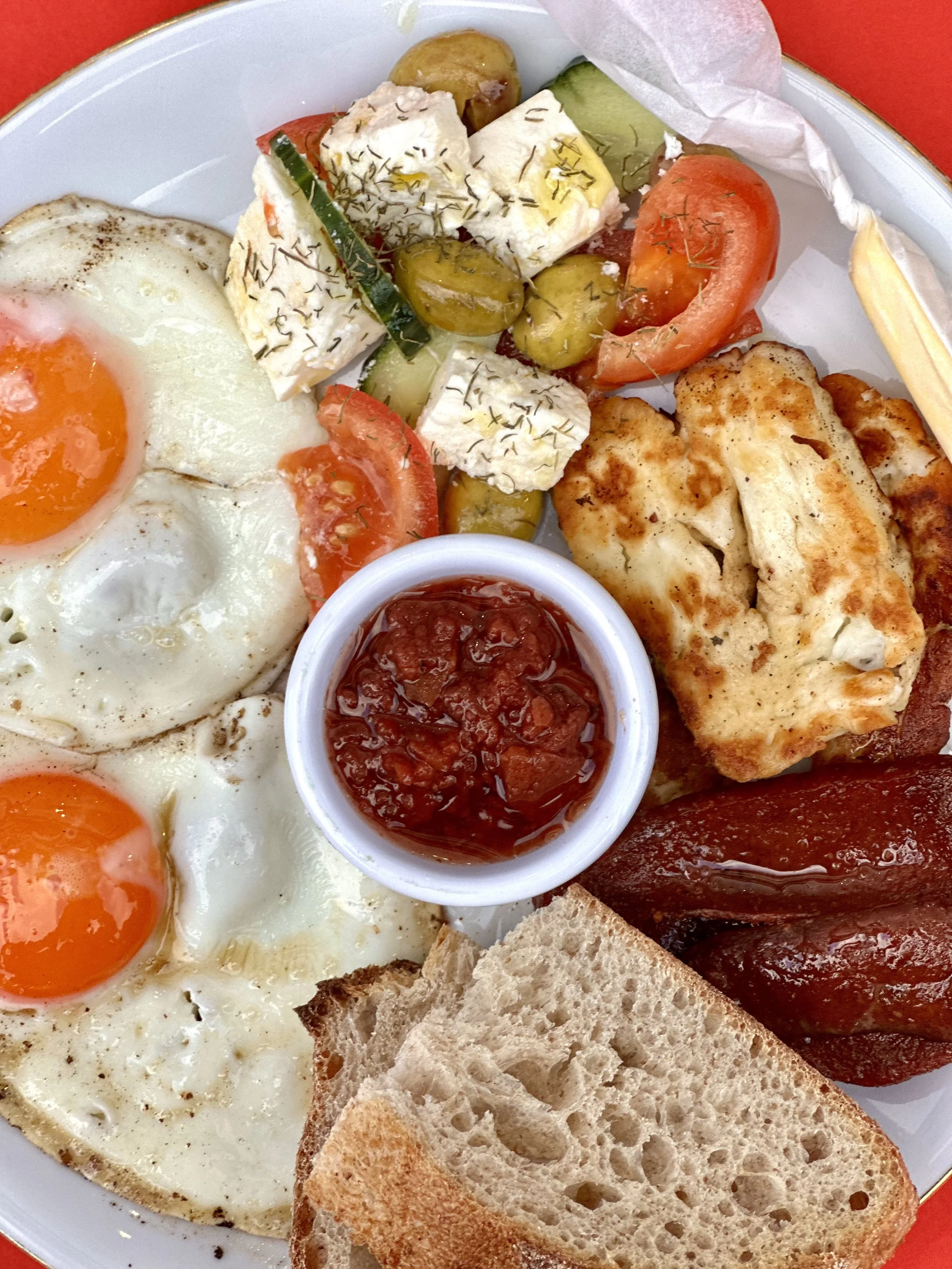 A plate of breakfast food including sunny side up eggs, sliced bread, cherry tomatoes, cucumbers, cheese with herbs, green olives, a small container of ketchup, and a piece of grilled meat.