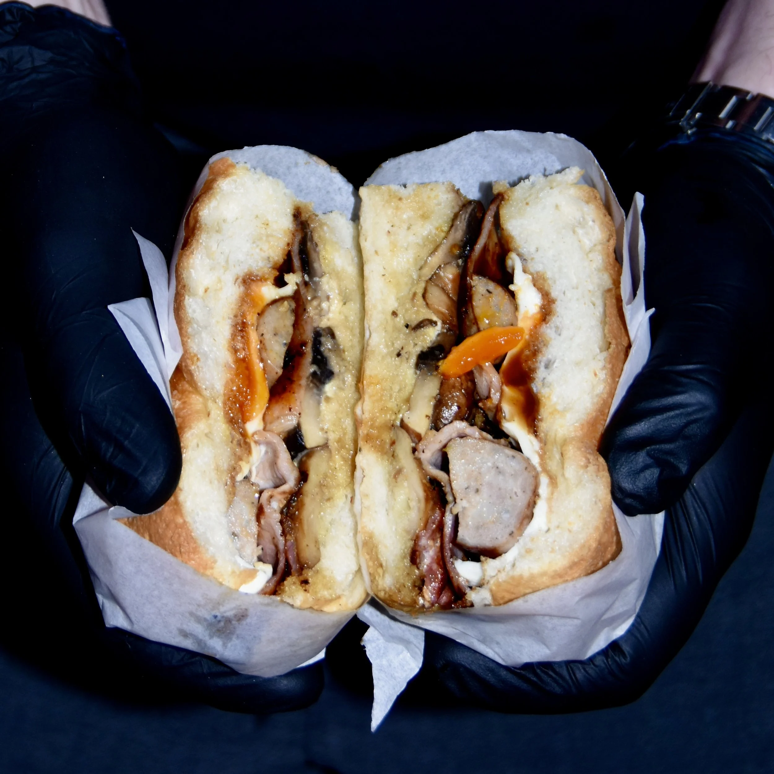 Half of a sandwich with cheese, mushrooms, and orange peppers on white bread, held by person wearing black gloves.
