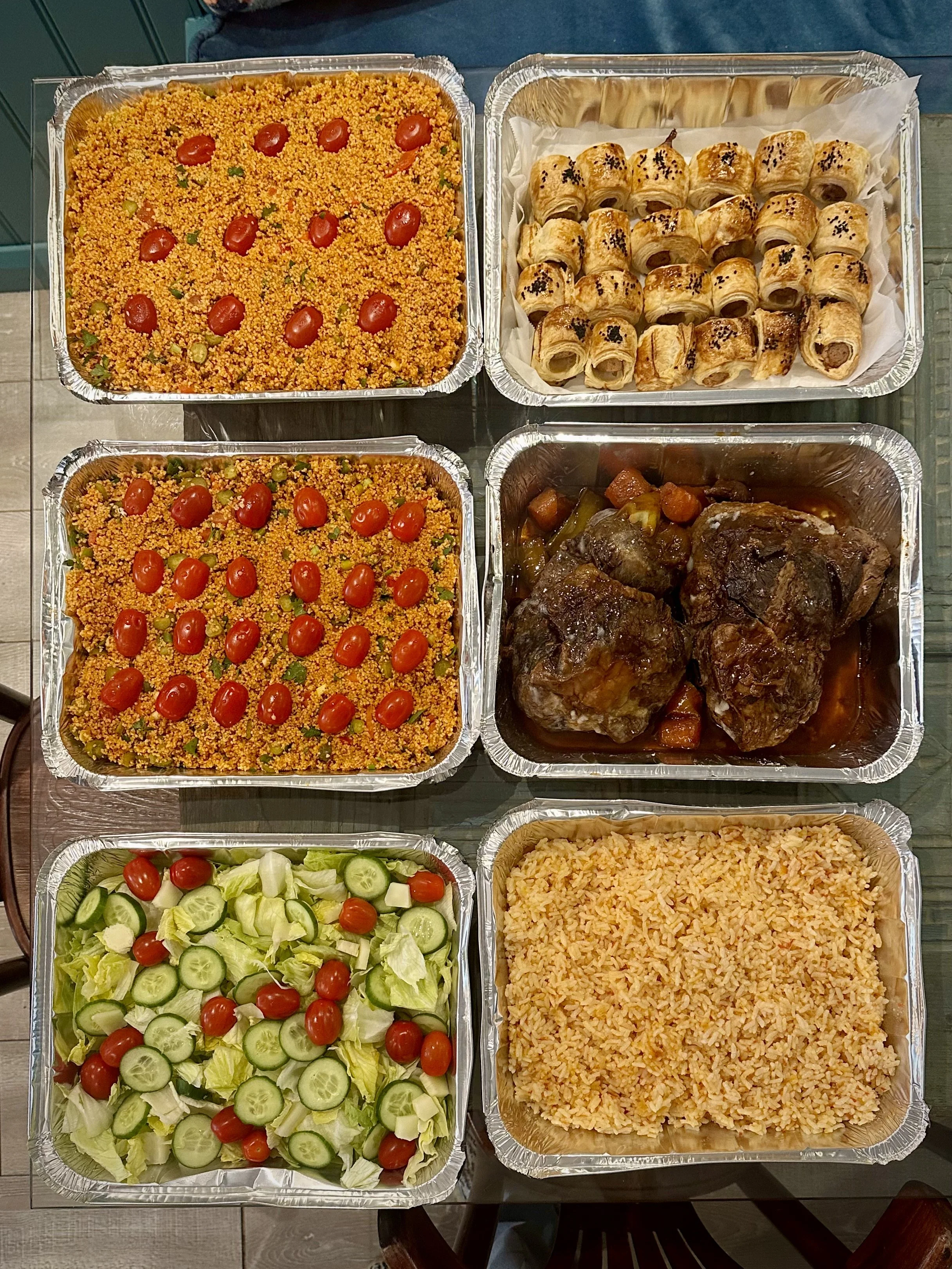 Various containers of food including salad, seasoned rice, roasted meat with vegetables, pastry rolls, and a side dish, all in aluminum trays.