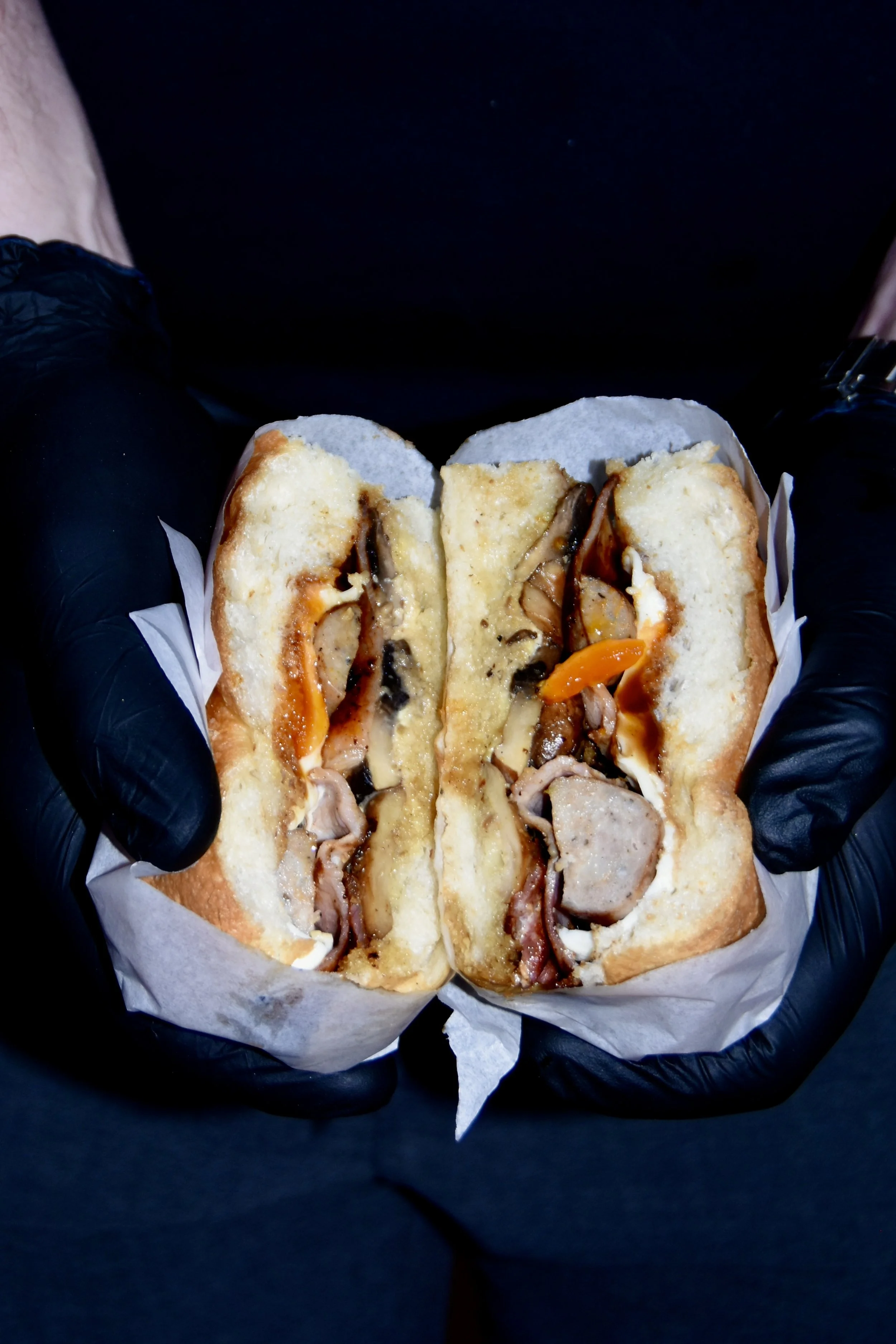 A person wearing black gloves holding a sandwich, cut in half, with visible layers of cheese, mushrooms, peppers, and possibly chicken or turkey on a bun, on white paper.