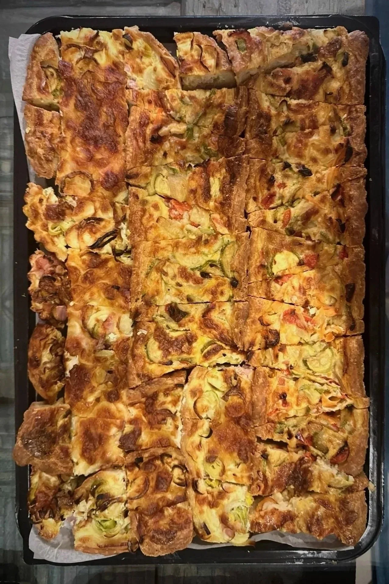 Large tray of assorted homemade pizza slices with various toppings including vegetables, cheese, and meats, baked and sliced into rectangles.