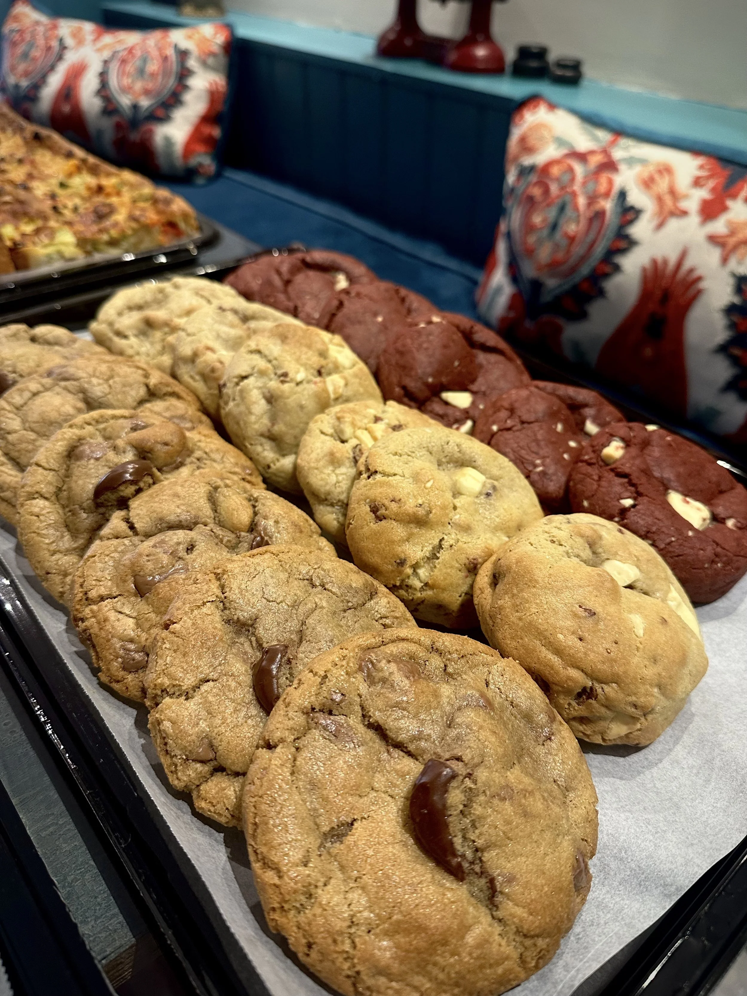 Display of assorted cookies, including chocolate chip and double chocolate chip, on a tray with patterned cushions in the background.