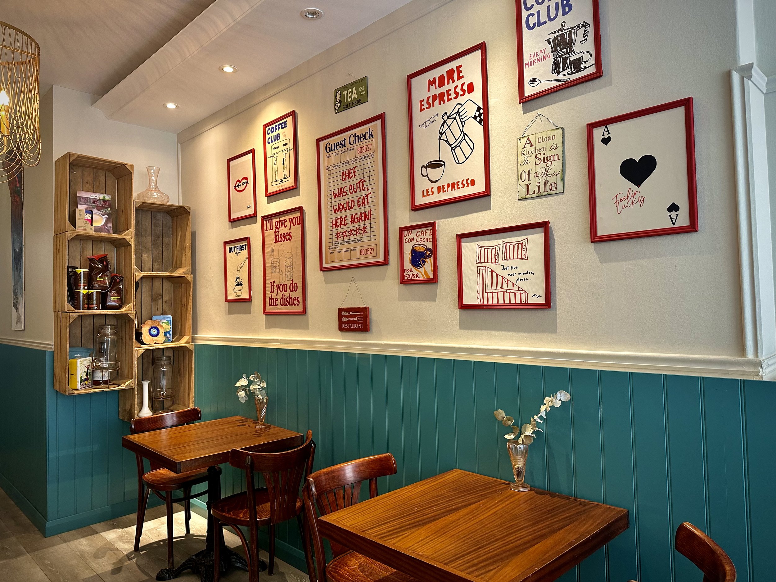Interior of a cozy coffee shop with teal wainscoting and cream walls, decorated with framed signs, a wooden shelving unit with coffee bags, and wooden tables with matching chairs.