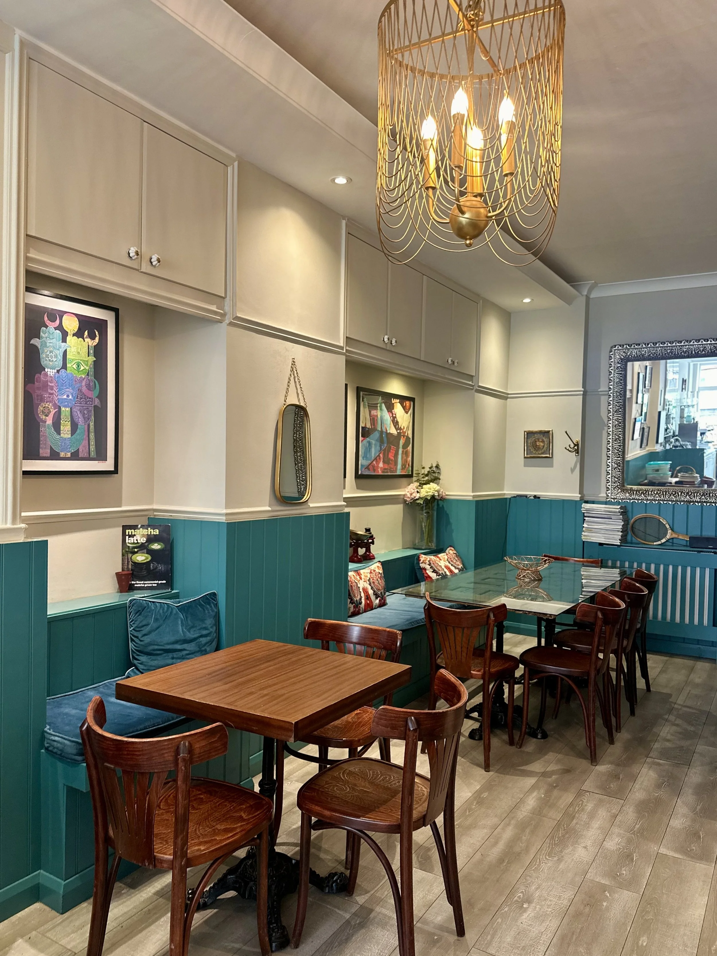 A cozy cafe interior with wooden tables and chairs, teal paneling along the walls, colorful framed artwork, decorative pillows on a built-in bench, and a gold chandelier hanging from the ceiling.