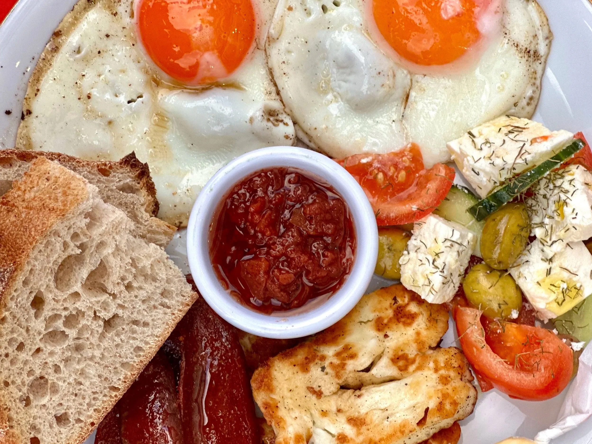A plate of breakfast food including two fried eggs with runny yolks, toasted bread, tomato slices, feta cheese with herbs, roasted asparagus with herbs, and a small cup of tomato-based sauce or jam.