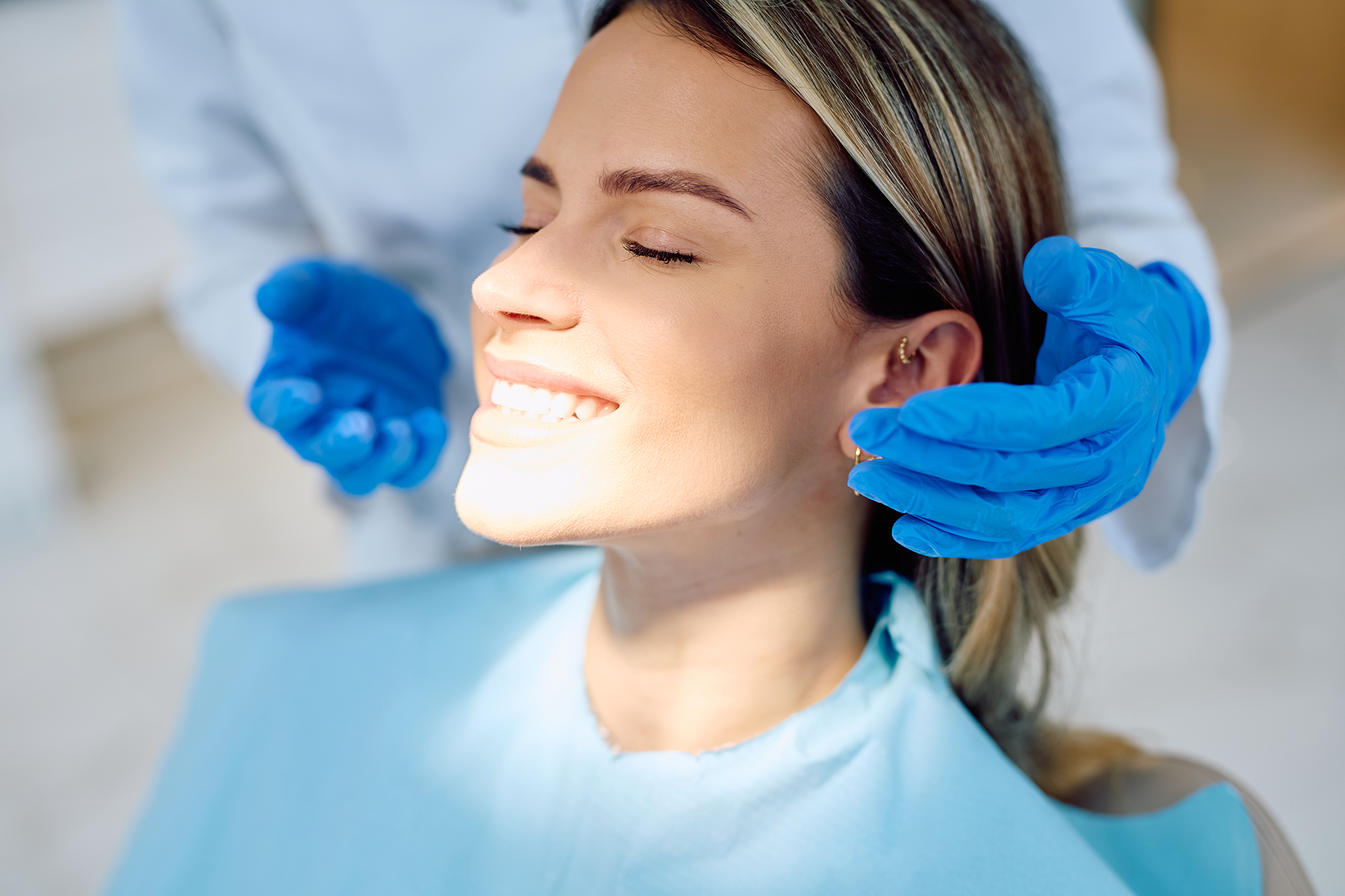 Kvinnelig pasient i tannlegestol som smiler mens to hender med blå hansker er på hver sin side av hodet