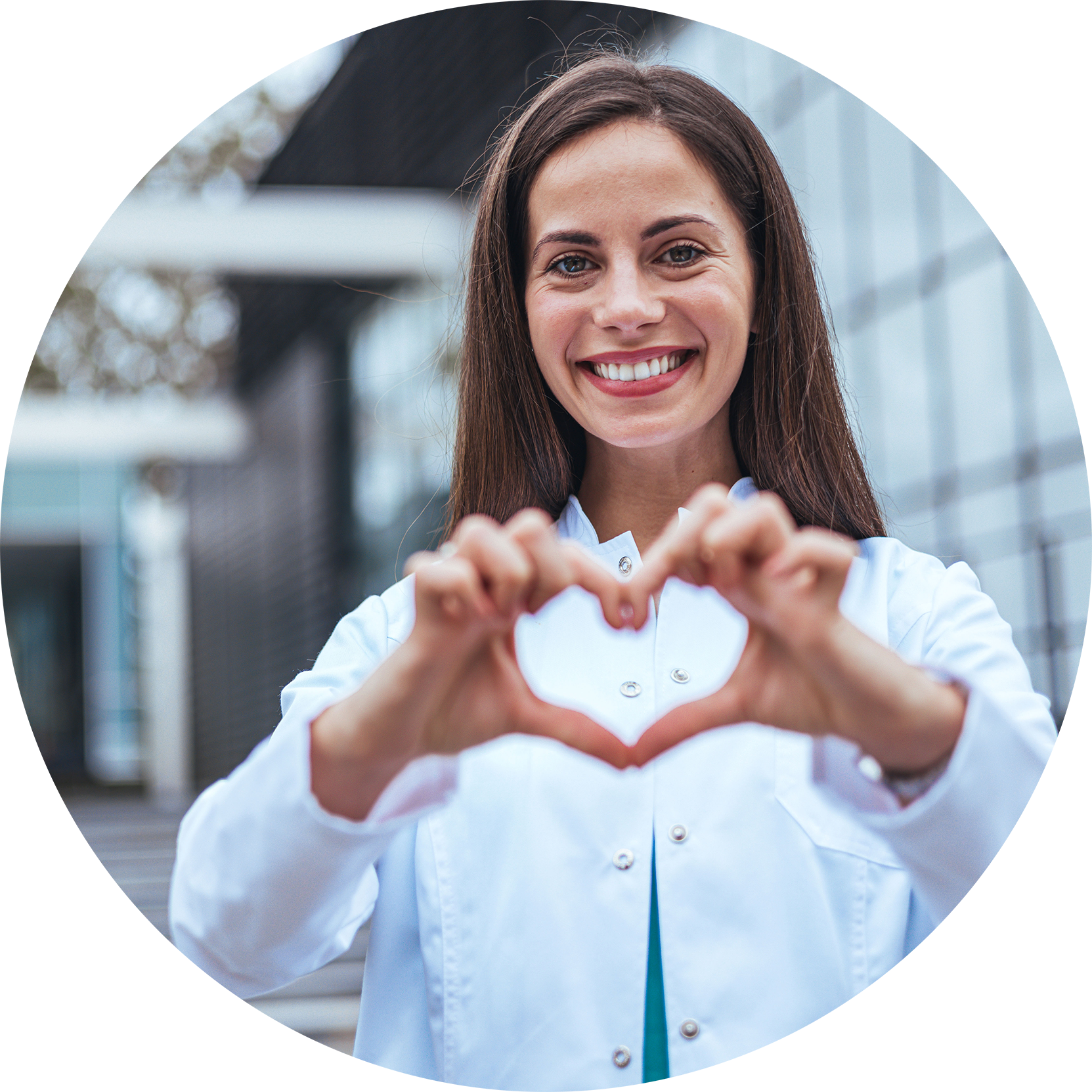 A young woman in a white coat smiling and making a heart with her hands in front of her, standing outside near a modern building with glass walls.