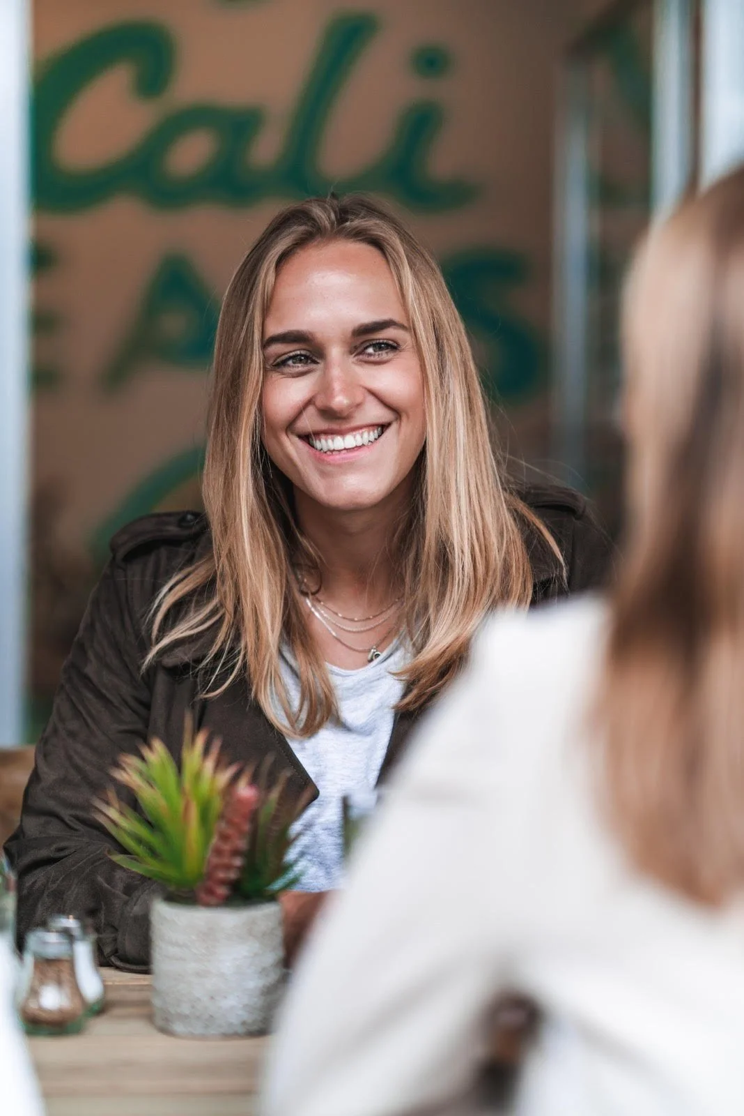 A woman with long blond hair smiling and sitting at a table in a cafe or restaurant, with a potted plant on the table.