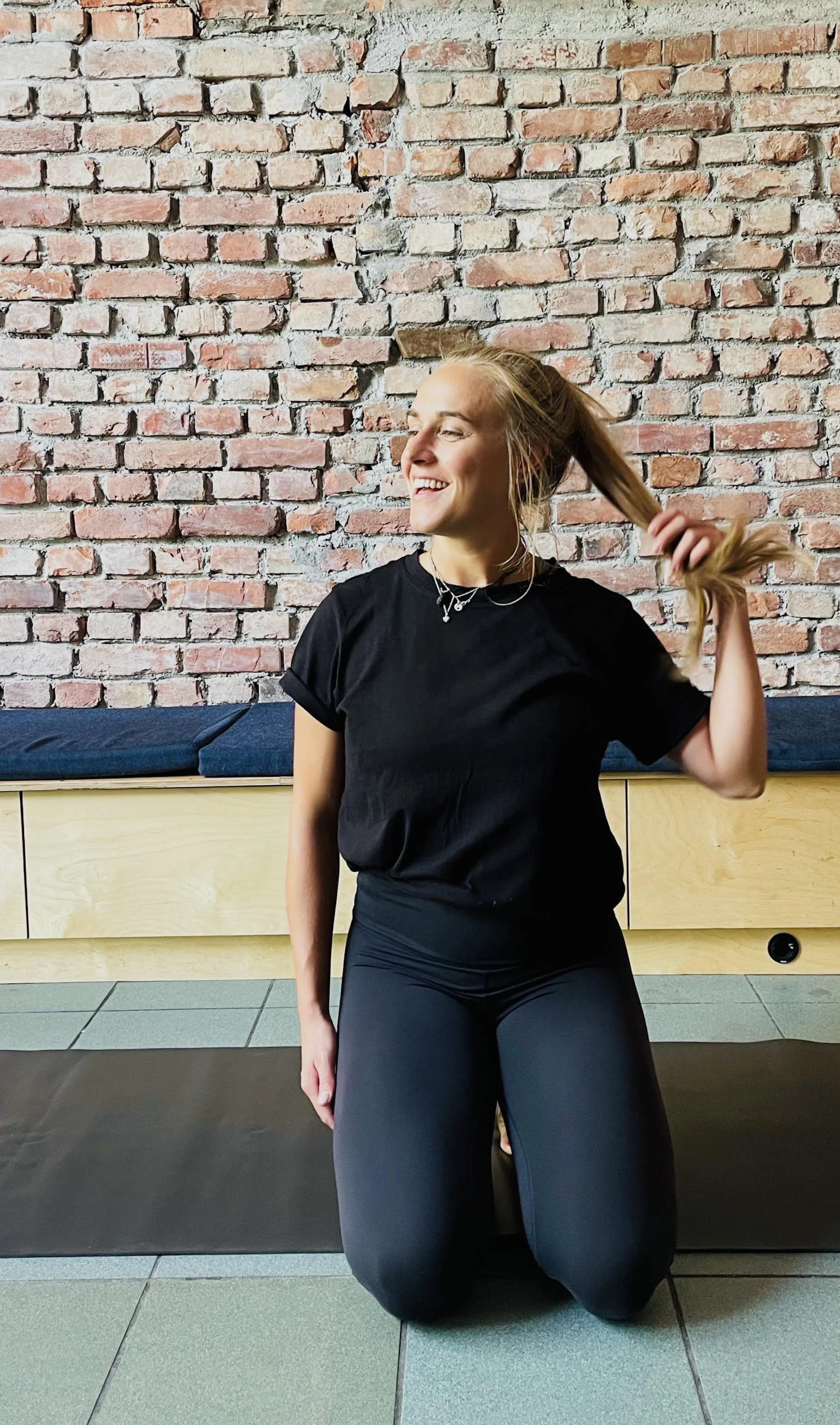 A young woman with blonde hair, wearing a black t-shirt and black leggings, smiling while sitting on her knees with one hand holding her hair back, in front of a rustic brick wall and a wooden bench with blue cushions.