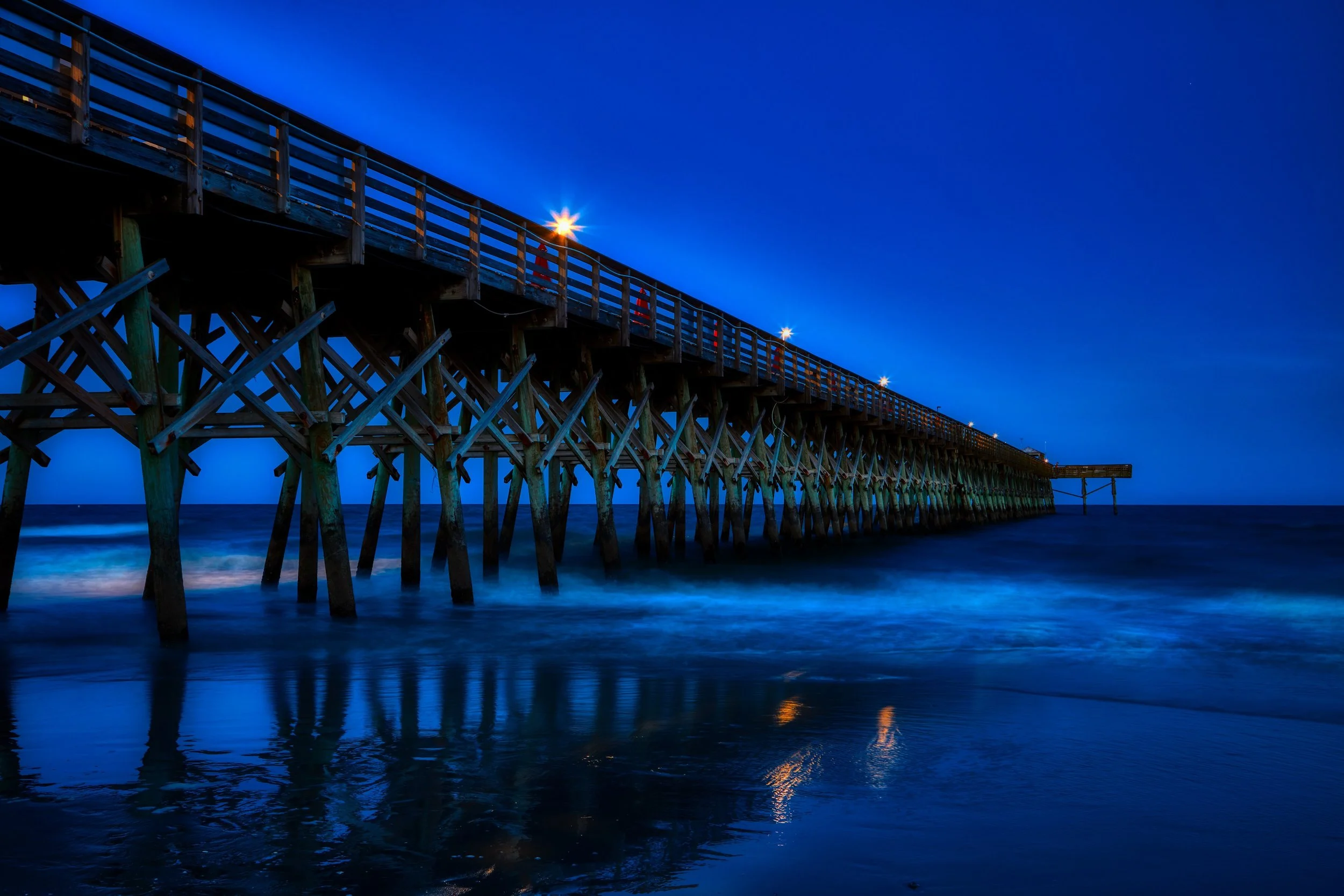 A wooden pier extends over the water at dusk, with bright lights illuminating the pier and their reflections visible on the water's surface.