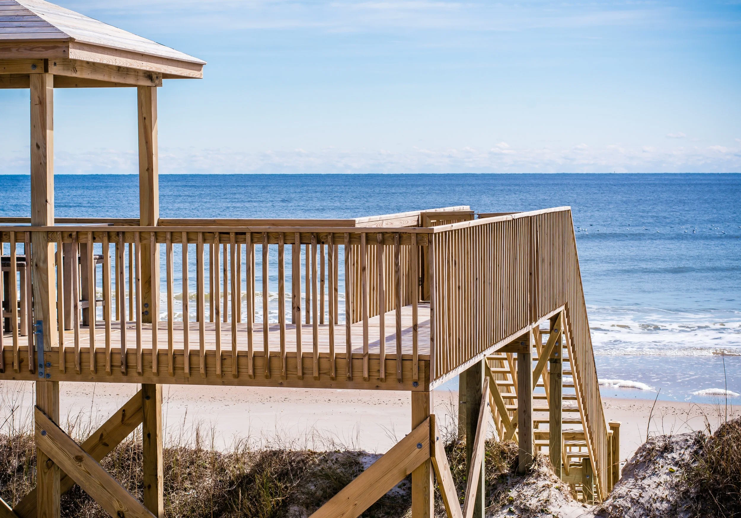 Wooden staircase and deck leading down to a sandy beach and the ocean with a blue sky and some clouds in the background.