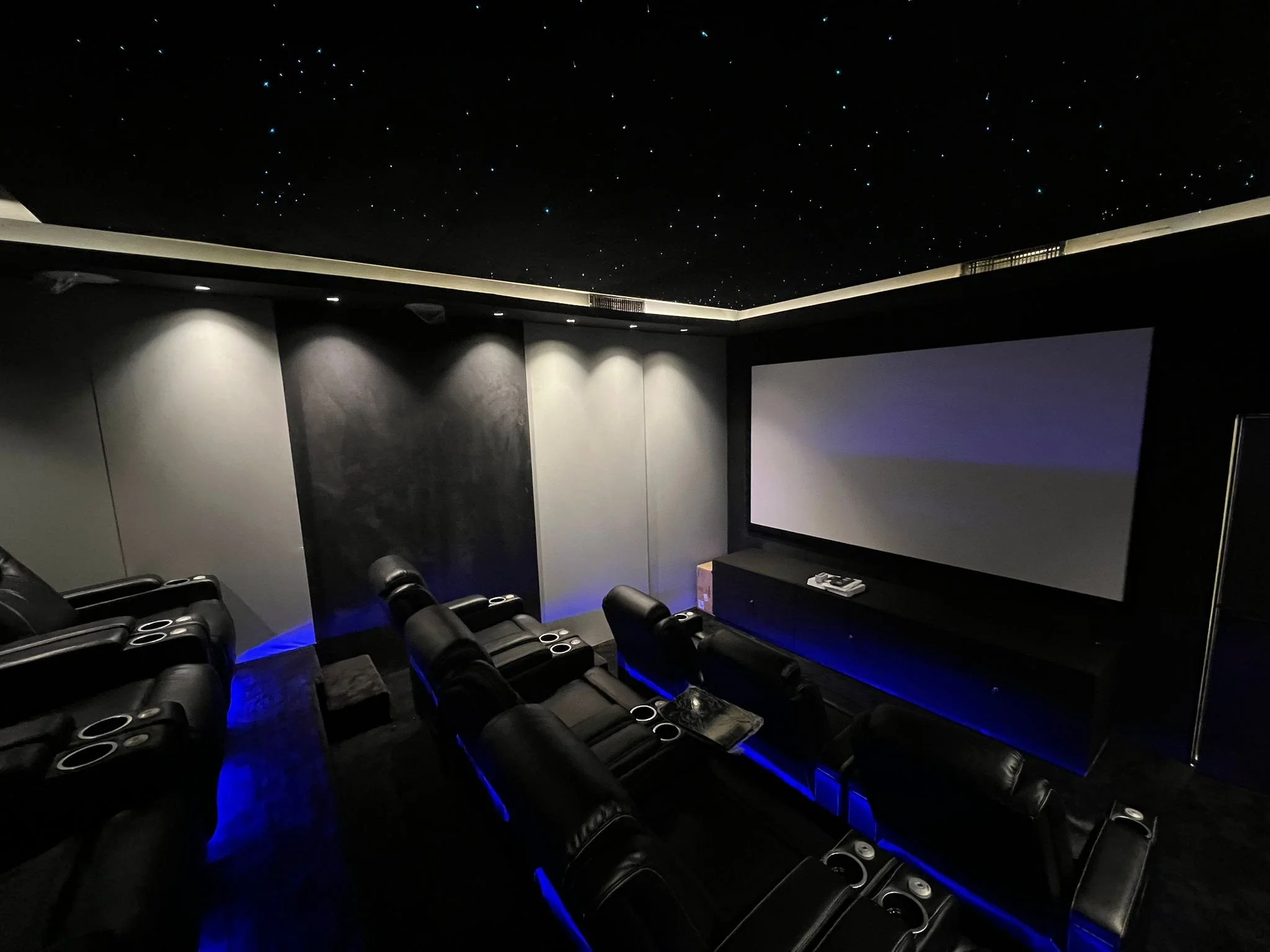 A dark movie theater with black leather recliner seats arranged in rows, illuminated by blue LED lighting along the aisle. The screen is blank, and the ceiling is decorated to resemble a starry night sky.