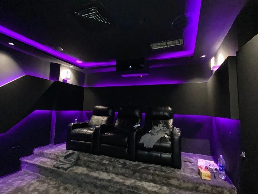 Home theater room with three black leather recliners on a carpeted platform, purple LED lighting along the walls and ceiling, with scattered items including water bottles, a box, and cloths.