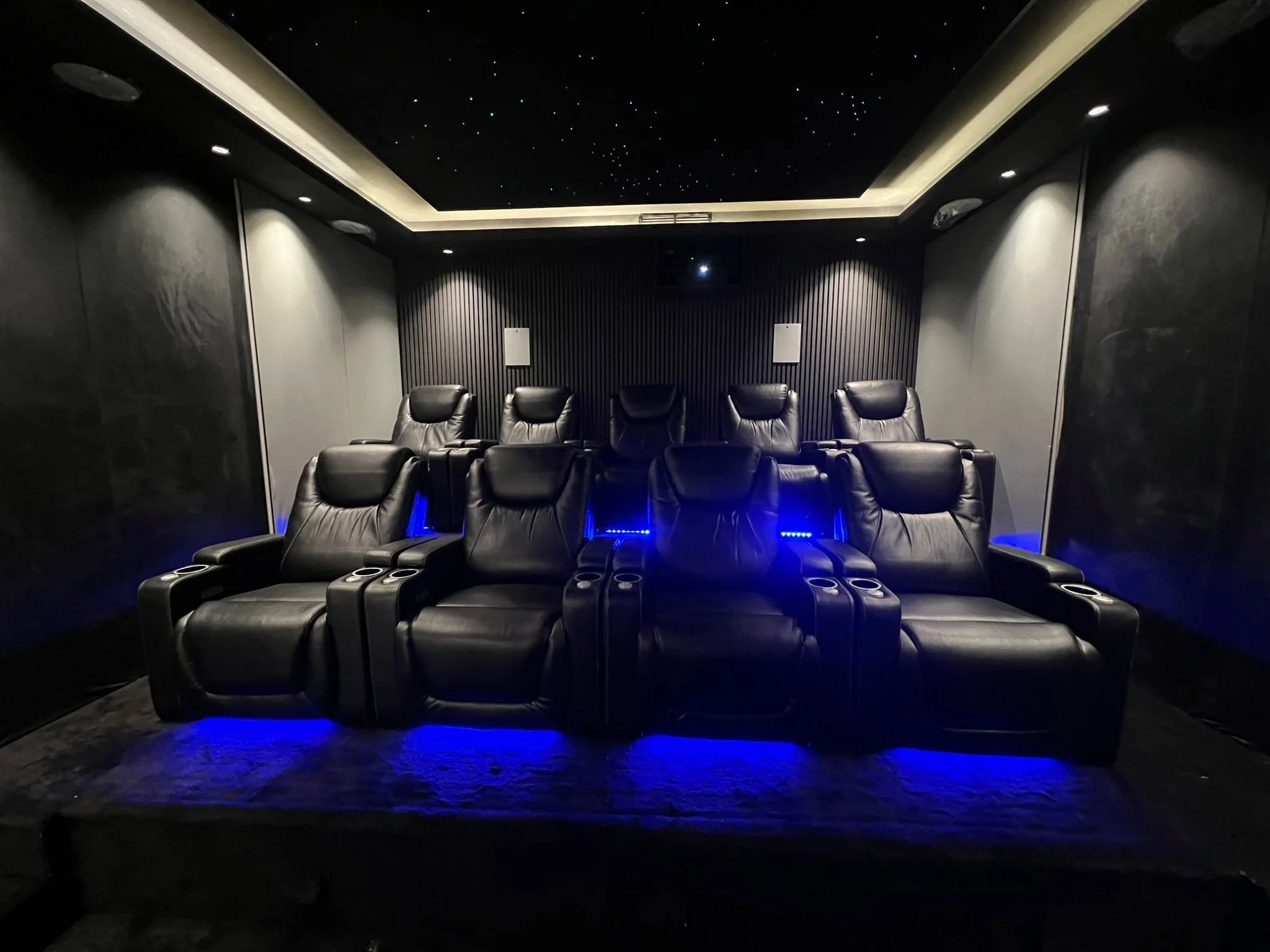 Home theater room with eight black leather recliner chairs arranged in two rows, modern black walls, starry ceiling, and blue LED lighting underneath the chairs.