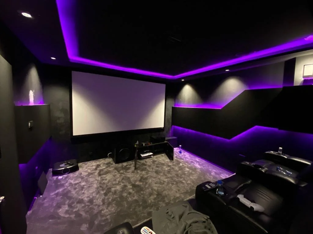 A home theater room with a large screen, black walls, black leather recliner, and purple LED accent lighting.