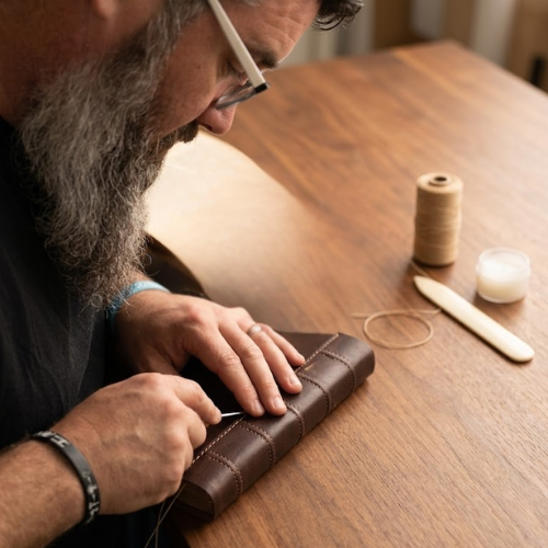 A man with glasses and a beard sewing a brown leather bible at a wooden table with thread, a needle, and sewing tools.