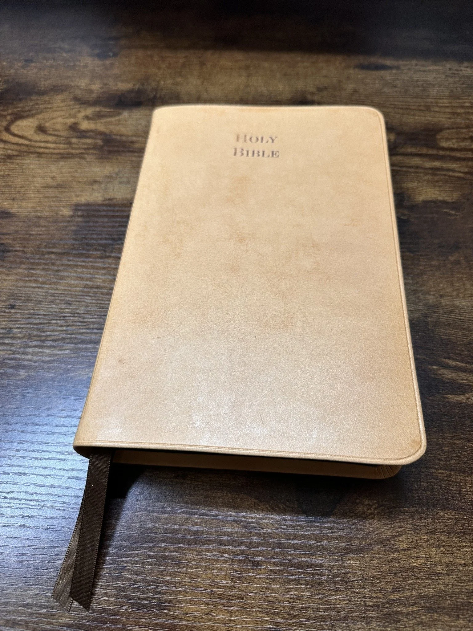 A closed, beige Holy Bible with a black ribbon bookmark lying on a dark wooden surface.