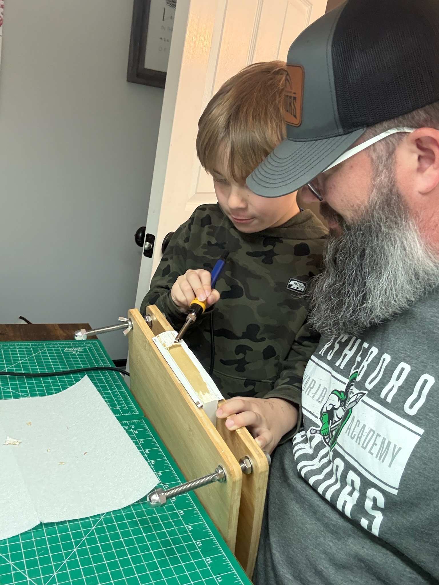 A young boy and an adult man working together on a bible rebinding project. The boy is using a tool on a bible inside a small wooden frame, while the man supervises. They are seated at a worktable with a green cutting mat.