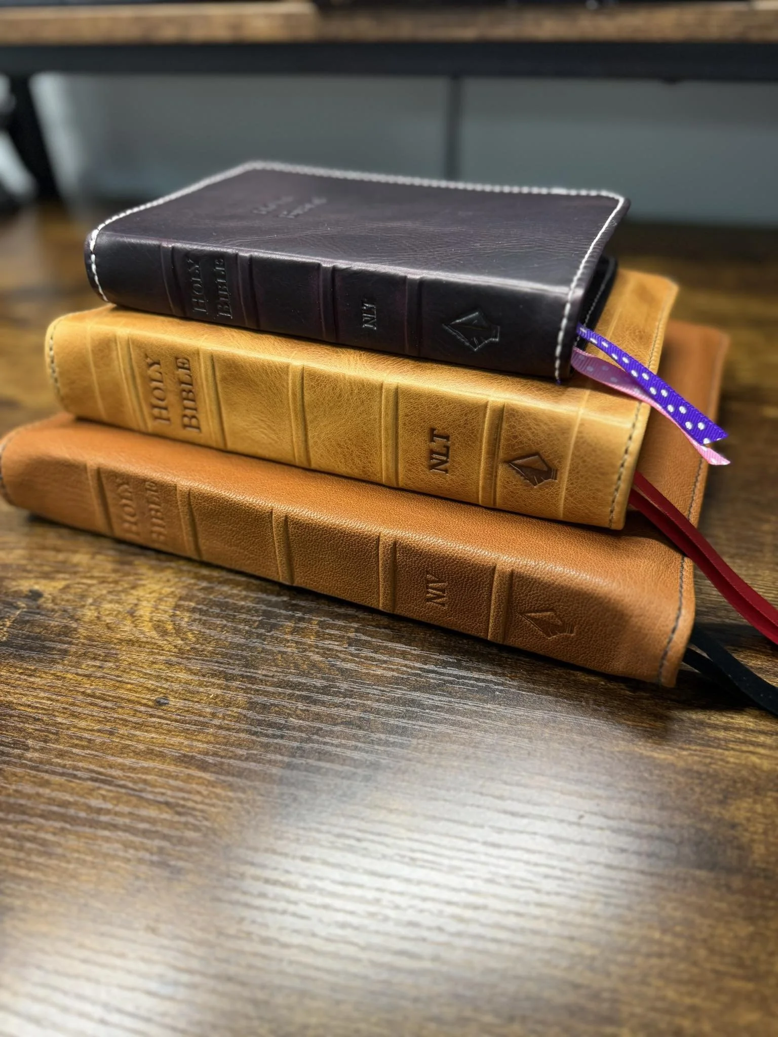 Three books stacked on a wooden surface, with the top book being a black Bible, the middle book a yellow Bible, and the bottom book an orange Bible.