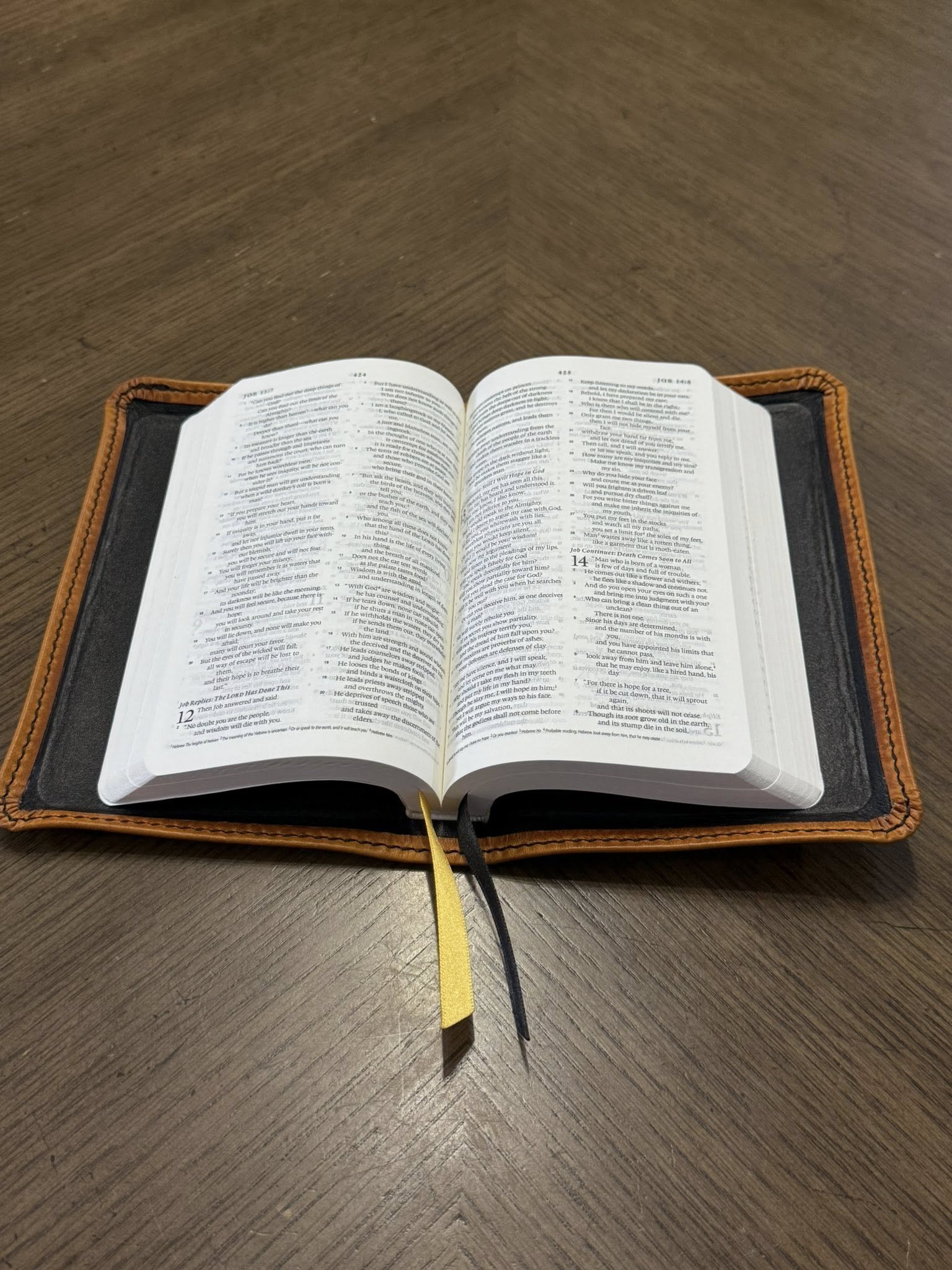 An open Bible on a wooden table with black and yellow ribbons marking pages, inside a black and orange leather cover.