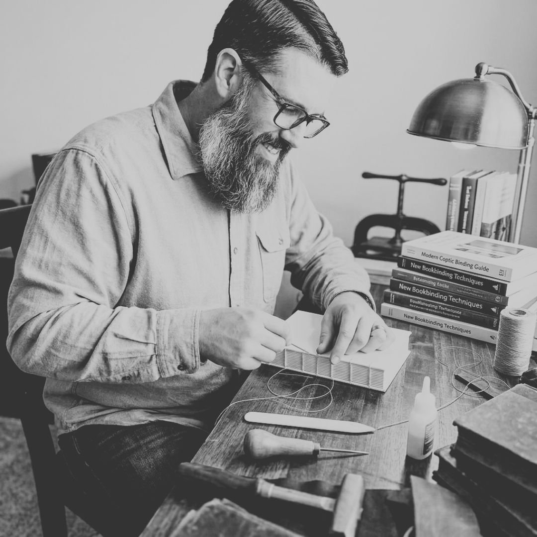 A man with glasses and a beard sitting at a desk, working on a small bookbinding project with tools and books on his workspace.