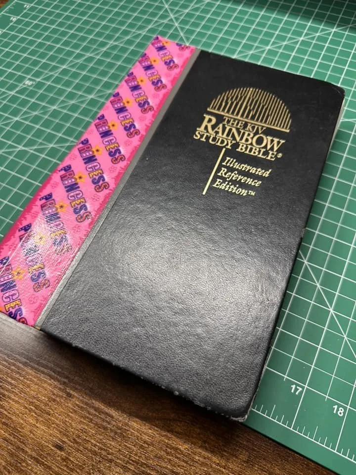 A black hardcover book titled "The New Rainbow Study Bible" resting on a green cutting mat with a pink ribbon placed alongside it.