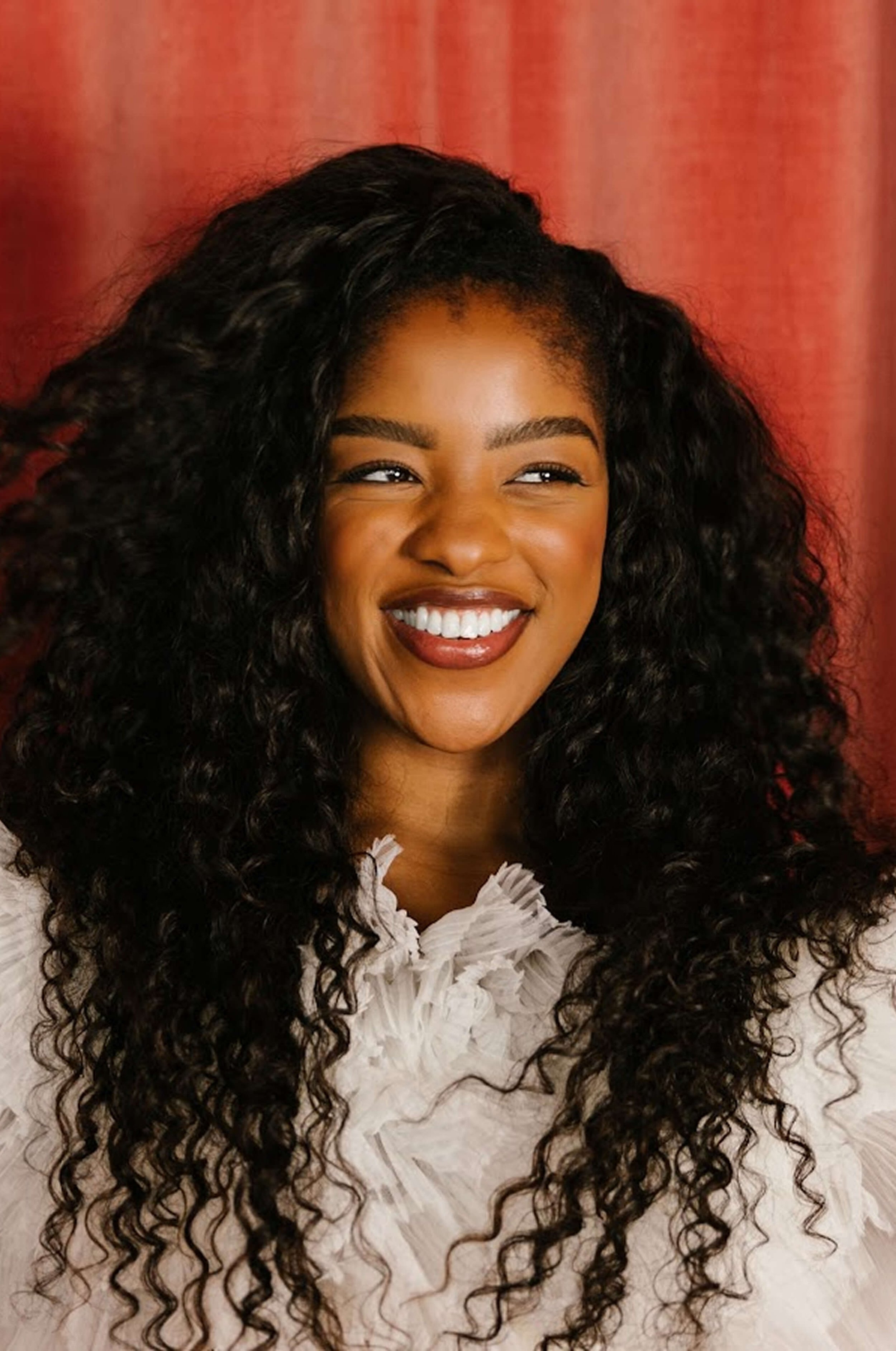 A smiling woman with long, curly black hair, wearing a white ruffled blouse, standing in front of a red curtain.