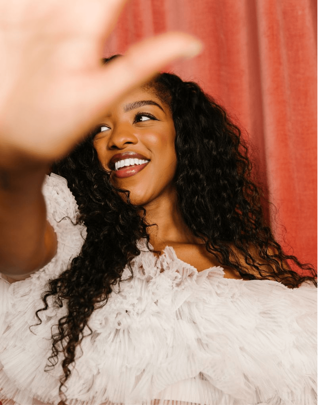 A woman with curly black hair, smiling and taking a selfie with a pinkish red curtain in the background.
