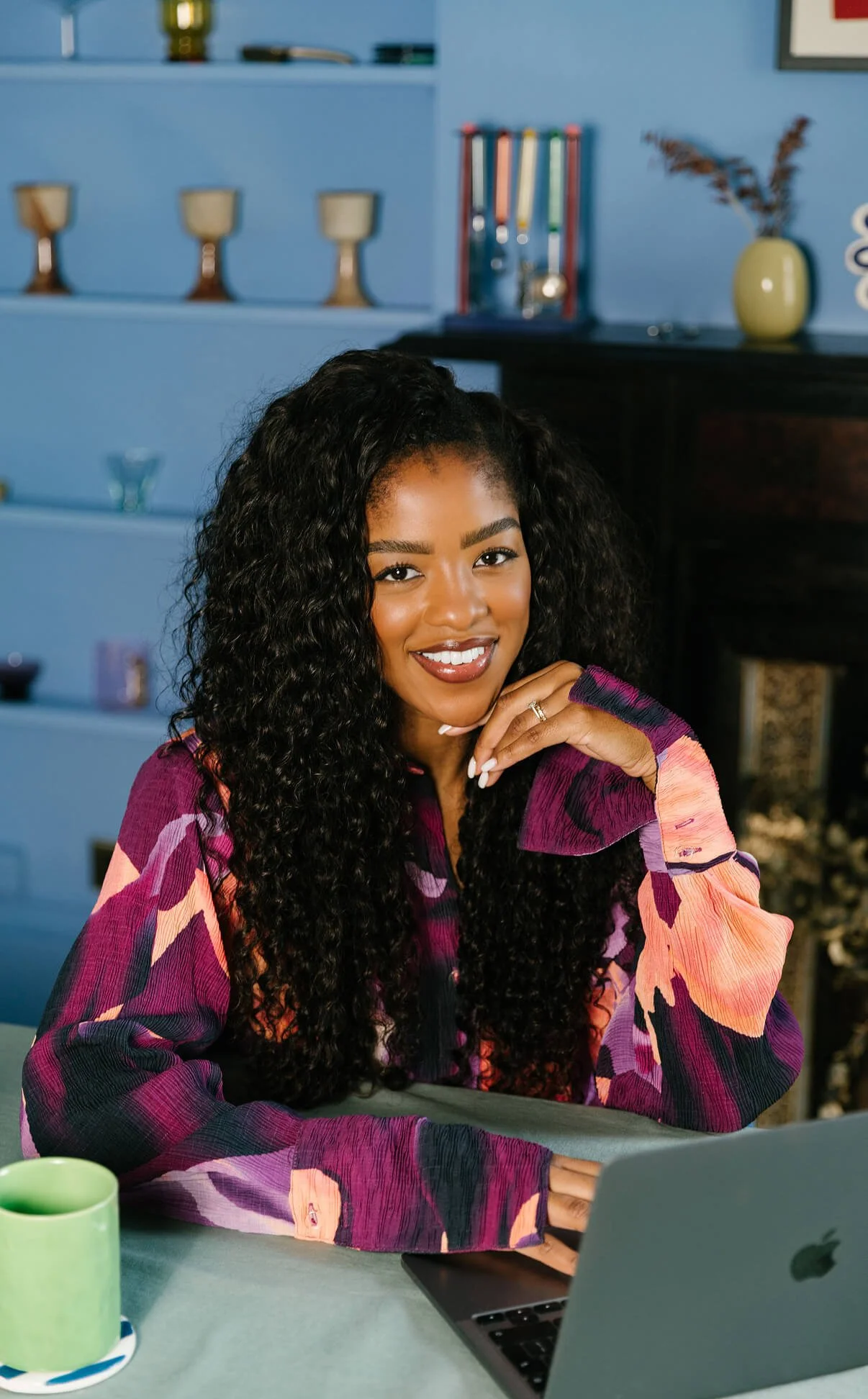 A woman with long curly black hair smiling at the camera, sitting at a table with a laptop and a green mug, in a room with blue walls and decorative items on shelves.