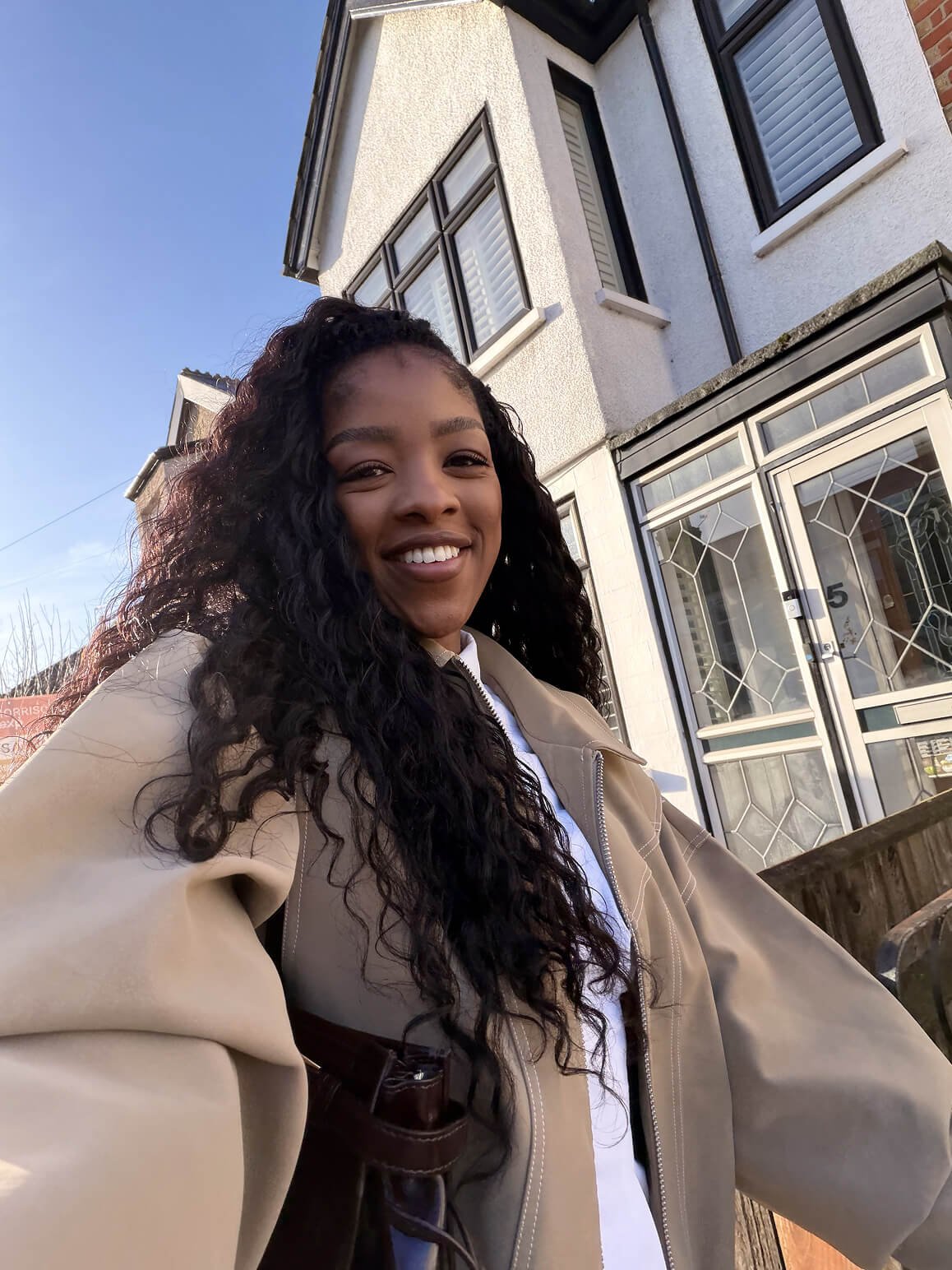 A woman with curly black hair smiling outdoors in front of a house with white siding and large windows, wearing a tan jacket.