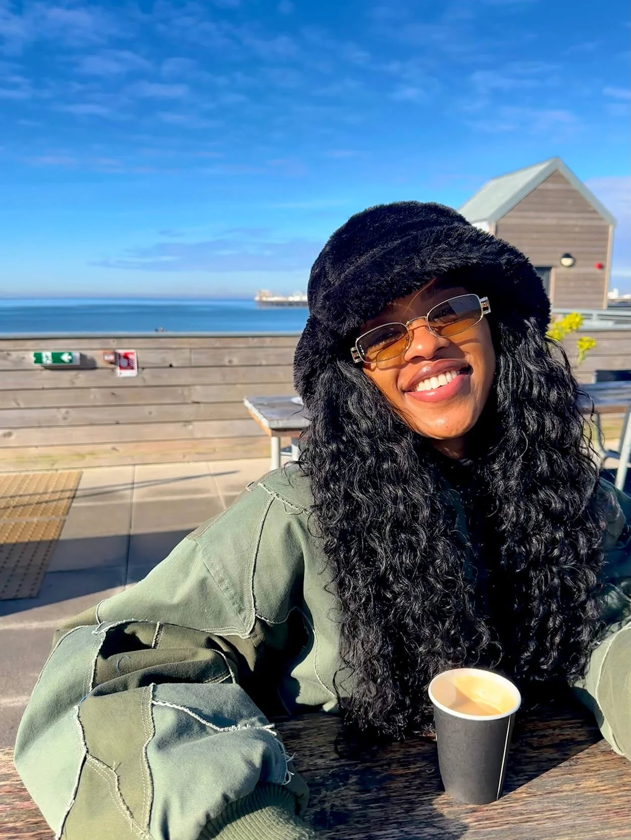 A woman with curly black hair smiling, wearing sunglasses, a black hat, and an olive-green jacket, sitting outdoors at a table with a small cup of coffee, overlooking the ocean with a blue sky and a wooden building in the background.