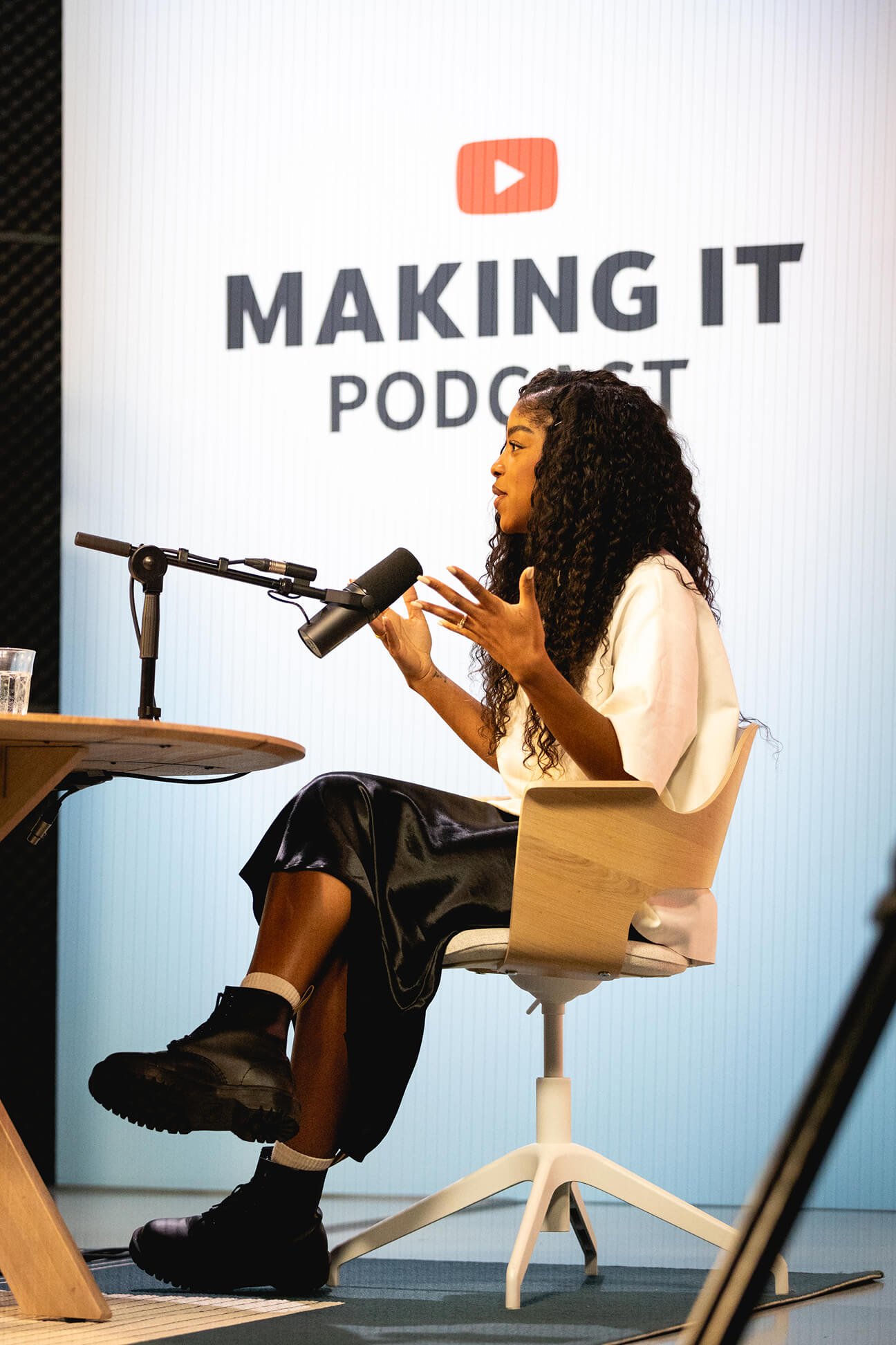 A woman with long curly hair talking into a microphone during a podcast recording session in front of a large screen displaying the YouTube logo and the words "Making It Podcast." She is sitting on a chair, wearing a white top and black skirt, and gesturing with her hands.