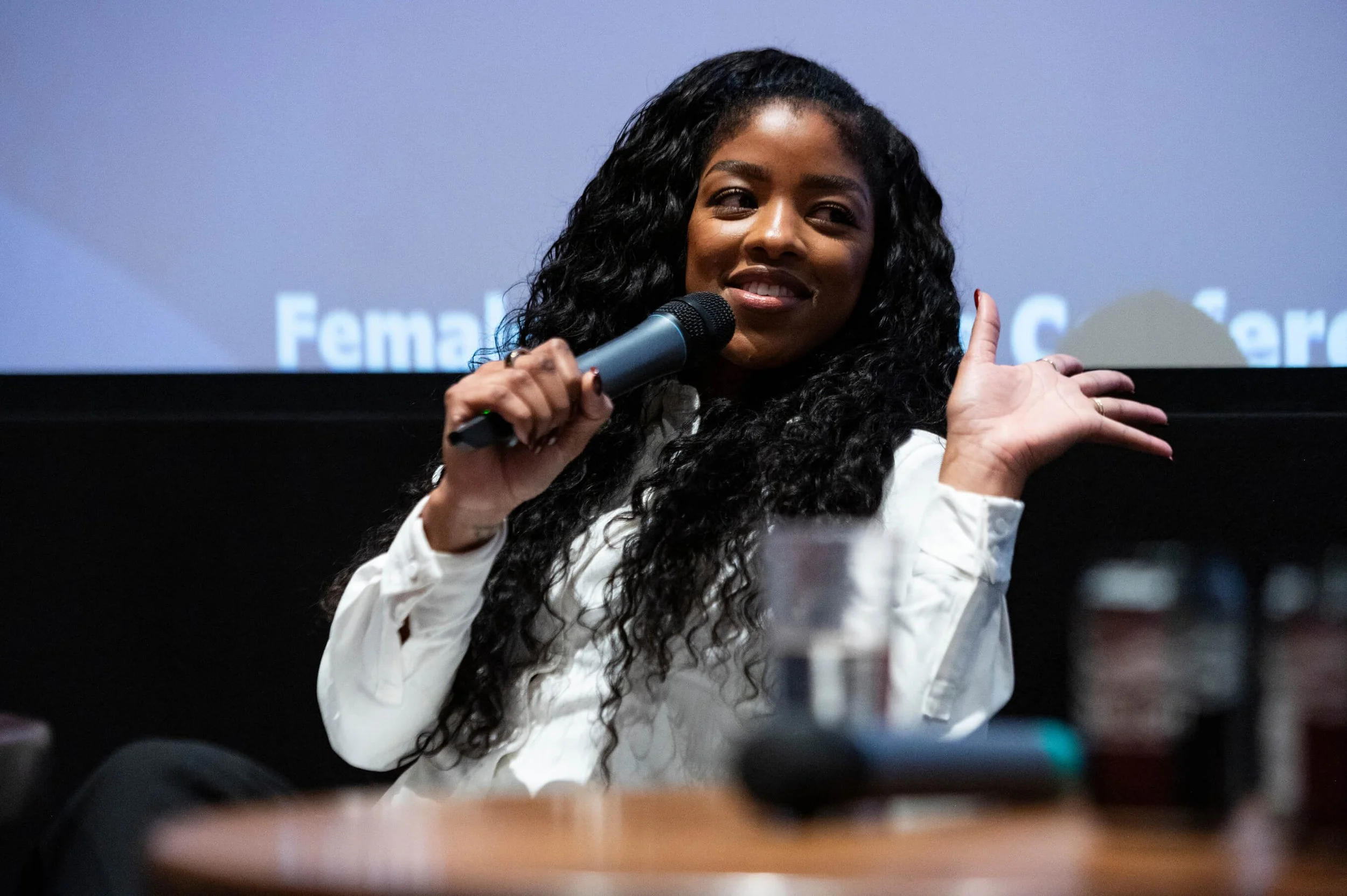 A woman with long curly black hair speaking into a microphone and gesturing with her right hand during a presentation.