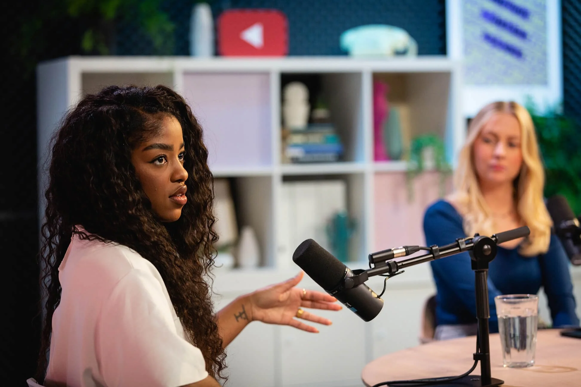 Two women sitting at a table with microphones, engaged in a discussion or interview, with bookcases and plants in the background.