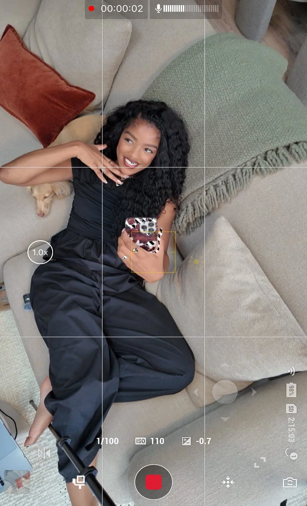 A woman with long curly black hair lying on a beige couch, smiling, holding a smartphone, next to a small light-colored dog, with decorative pillows around.
