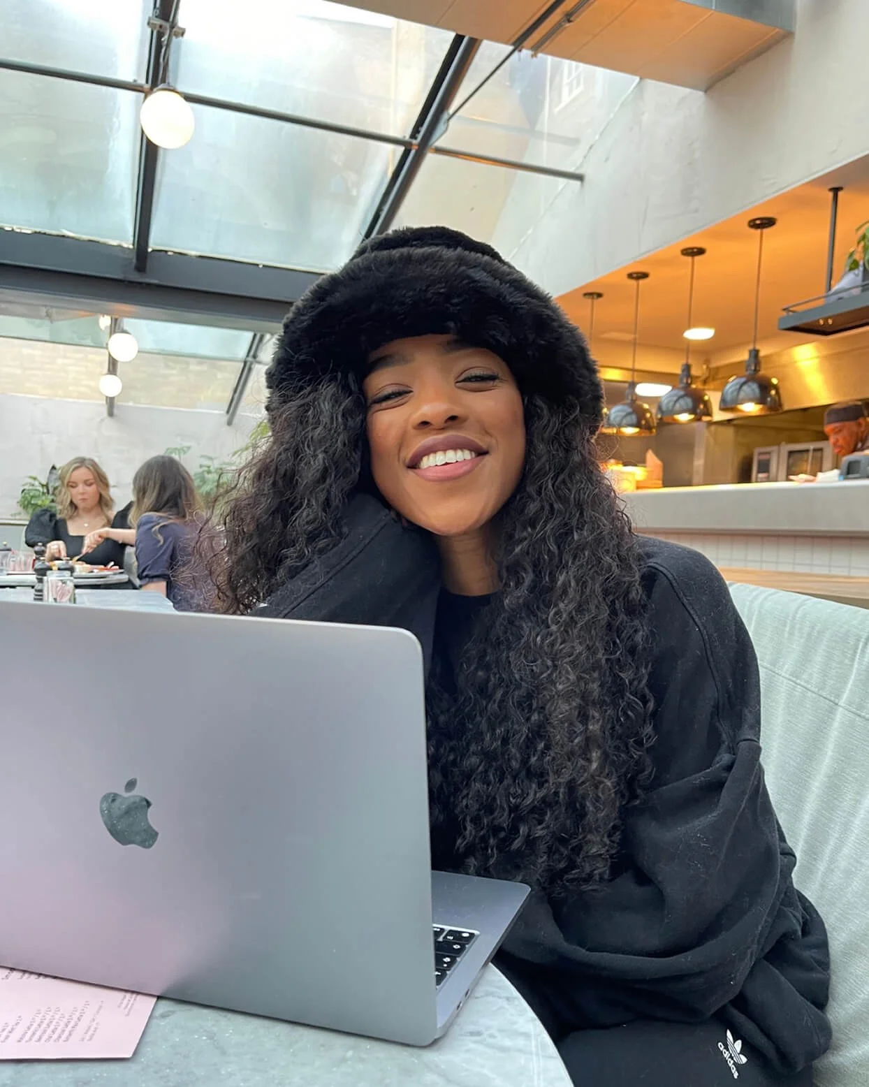 A smiling woman with long curly hair wearing a black fur hat and black jacket, sitting at a table with a silver laptop in a cafe.