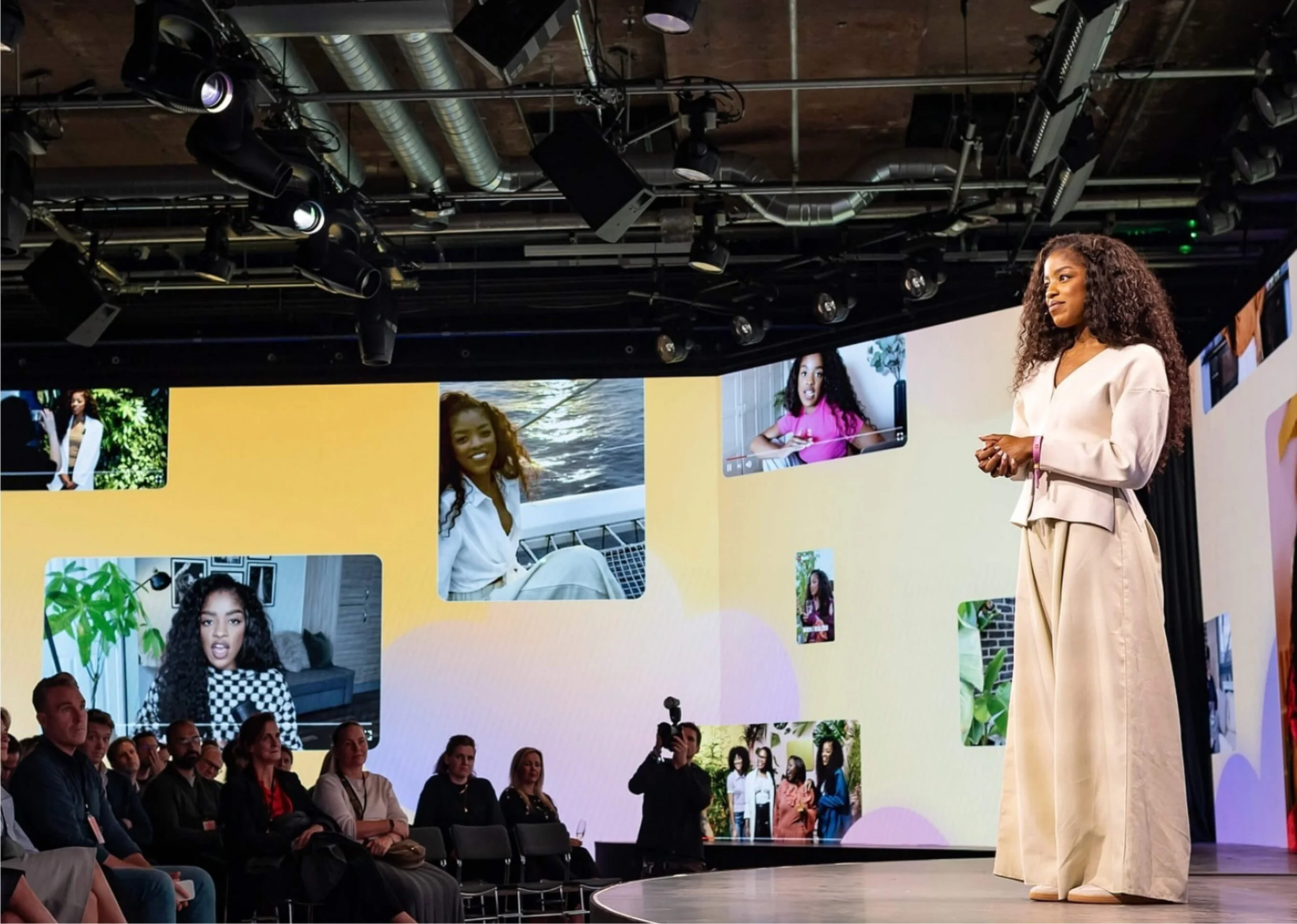 A woman with curly hair standing on stage speaking to an audience, with large screens behind her displaying images of diverse women in various settings.
