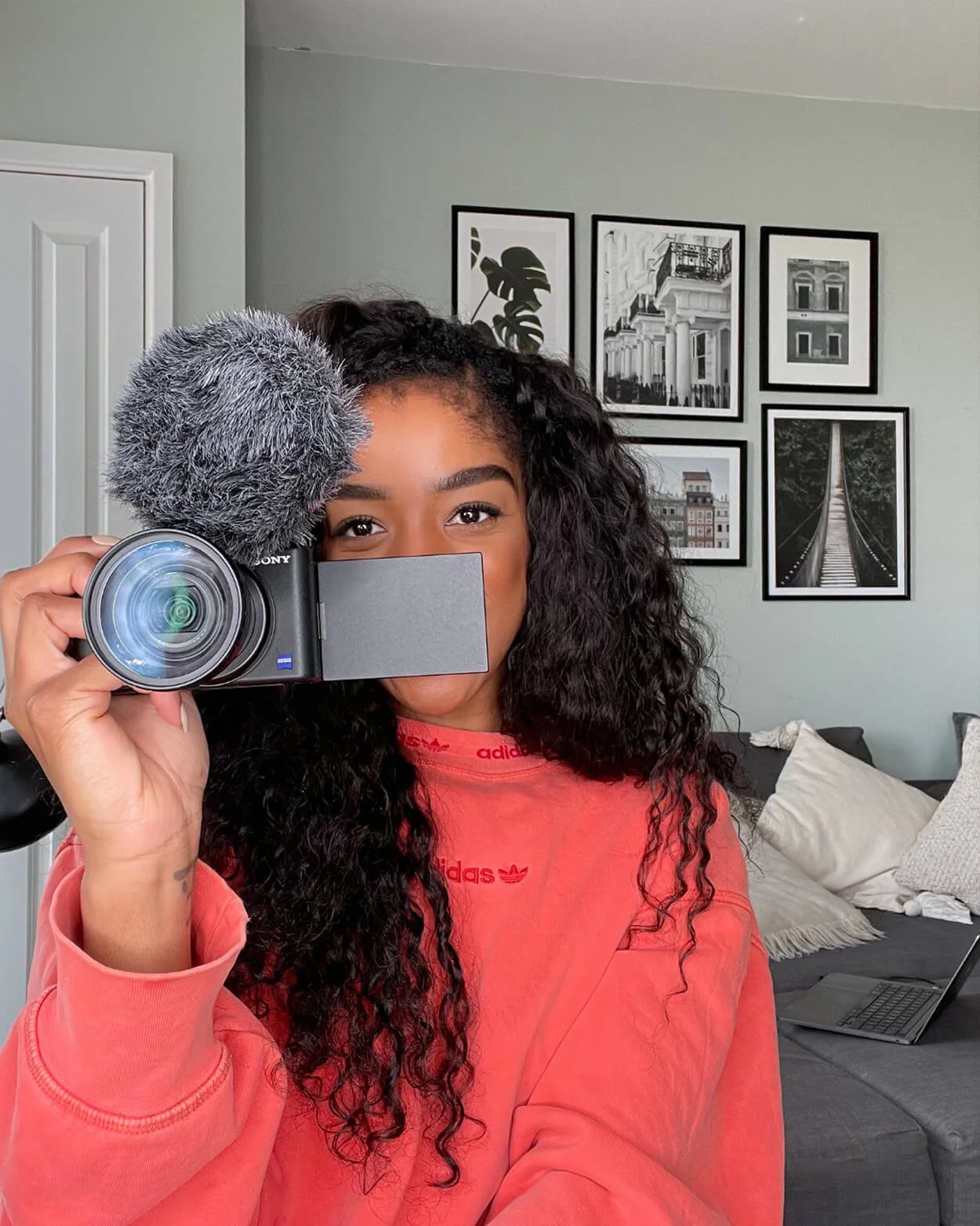 A young woman with long curly hair wearing a red Adidas sweatshirt, holding a camera with a microphone, taking a selfie in a living room with framed black and white pictures on the wall and a gray sofa with pillows.